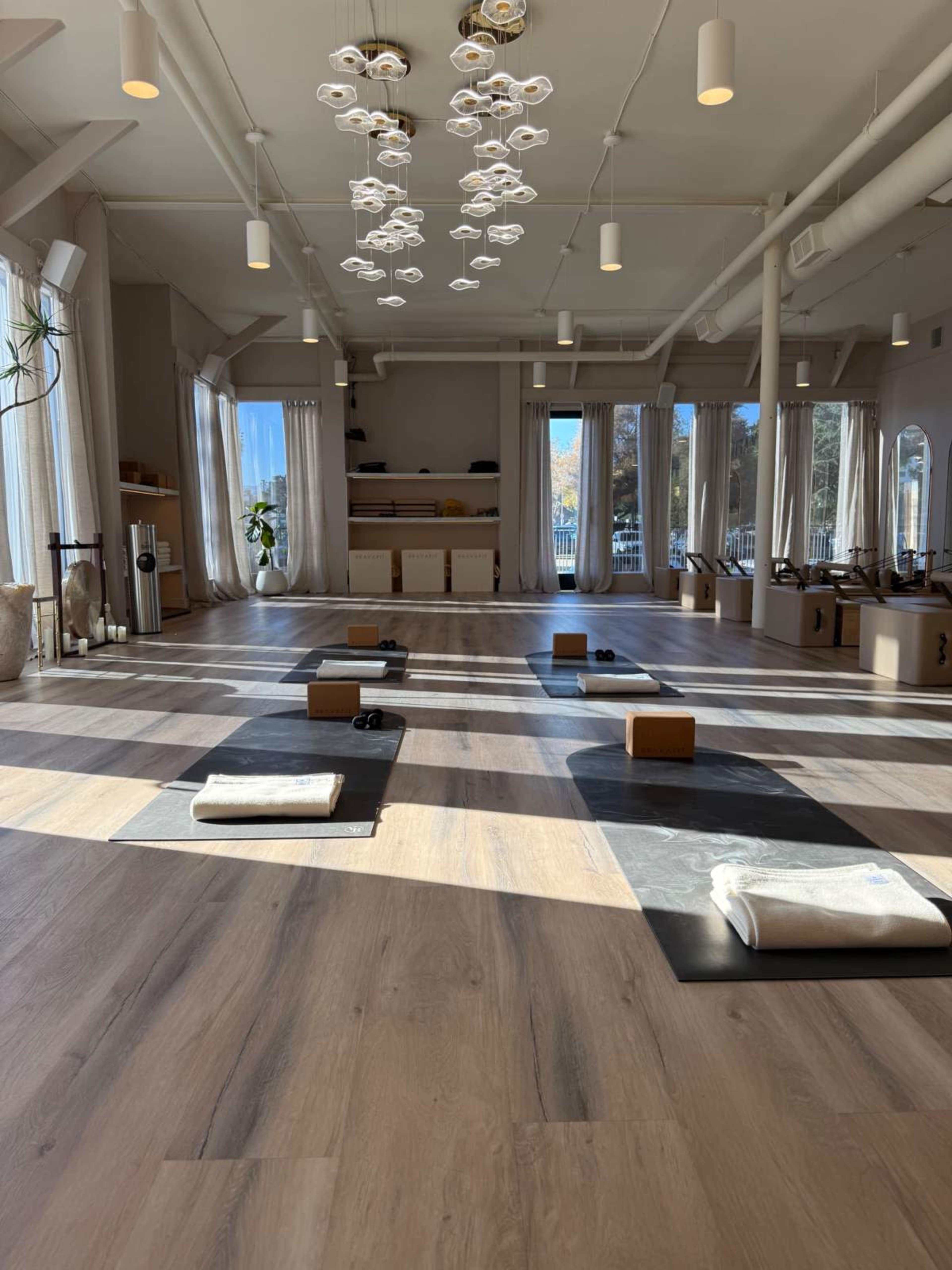 The image shows a spacious yoga studio with wooden flooring, multiple yoga mats arranged in rows, and large windows allowing natural light to illuminate the space.