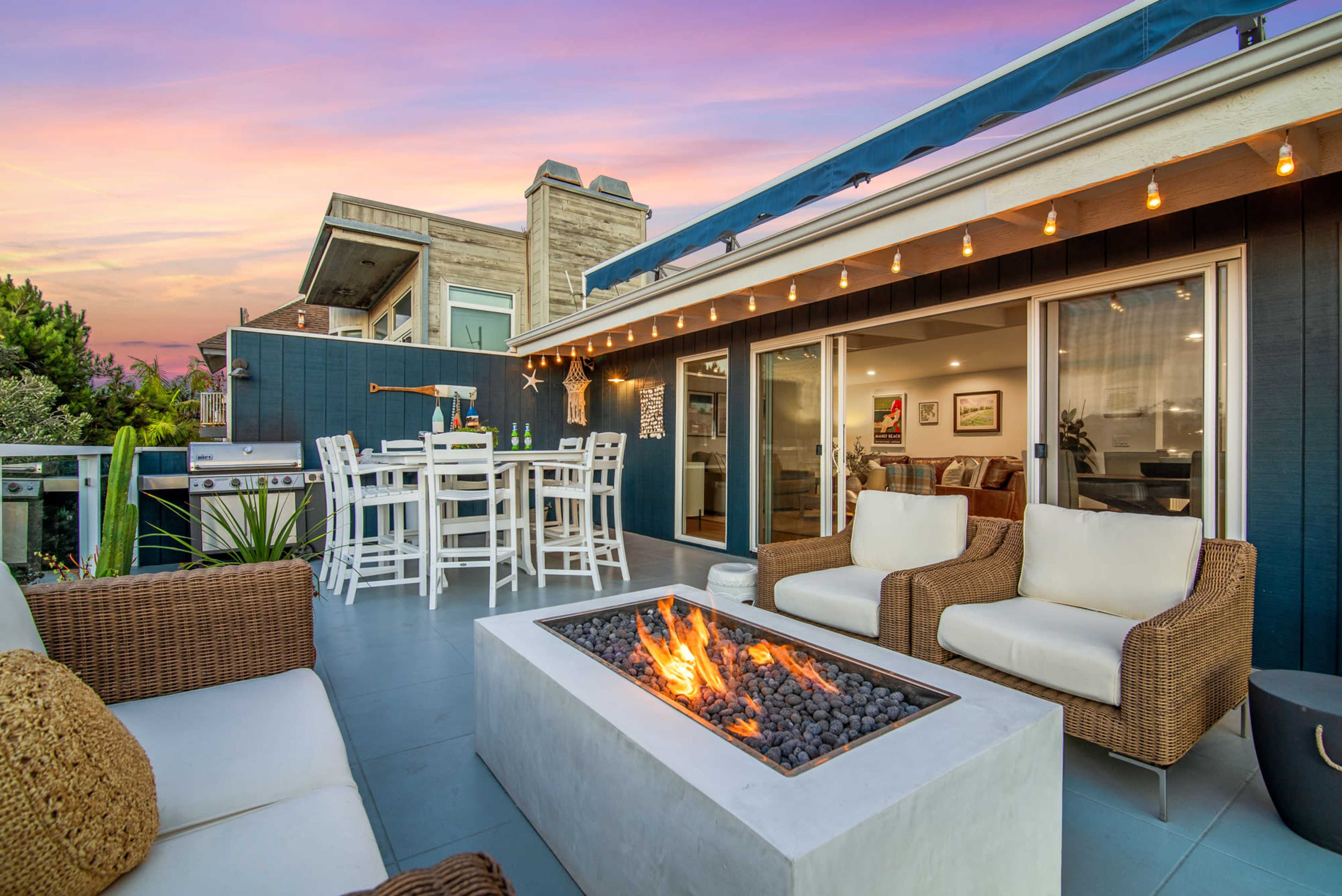 A residential patio area with a fire pit surrounded by seating, and a dining set nearby, with a sunset sky in the background.