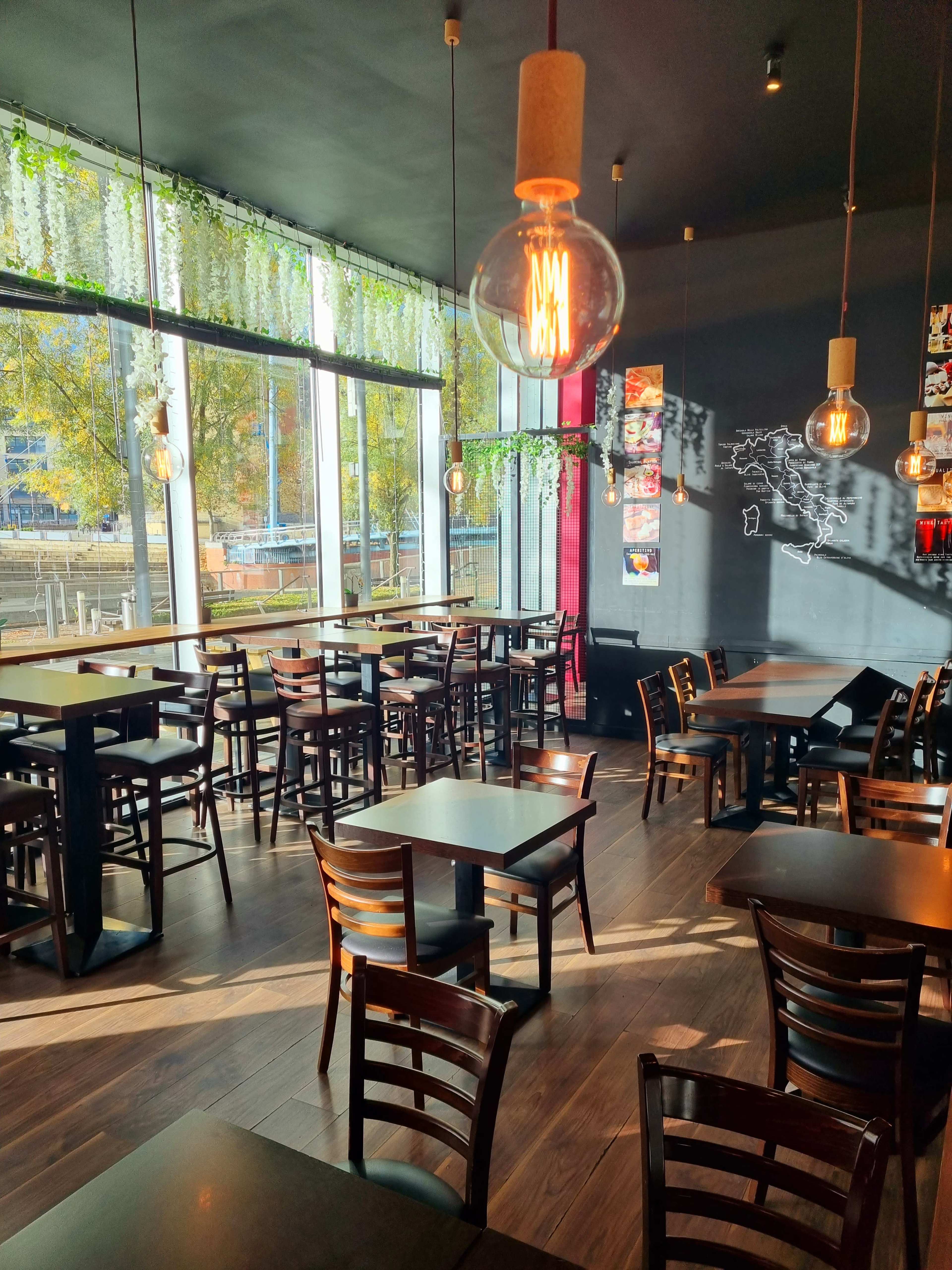 A modern restaurant interior with wooden tables and chairs, large windows providing natural light, and hanging pendant lights.