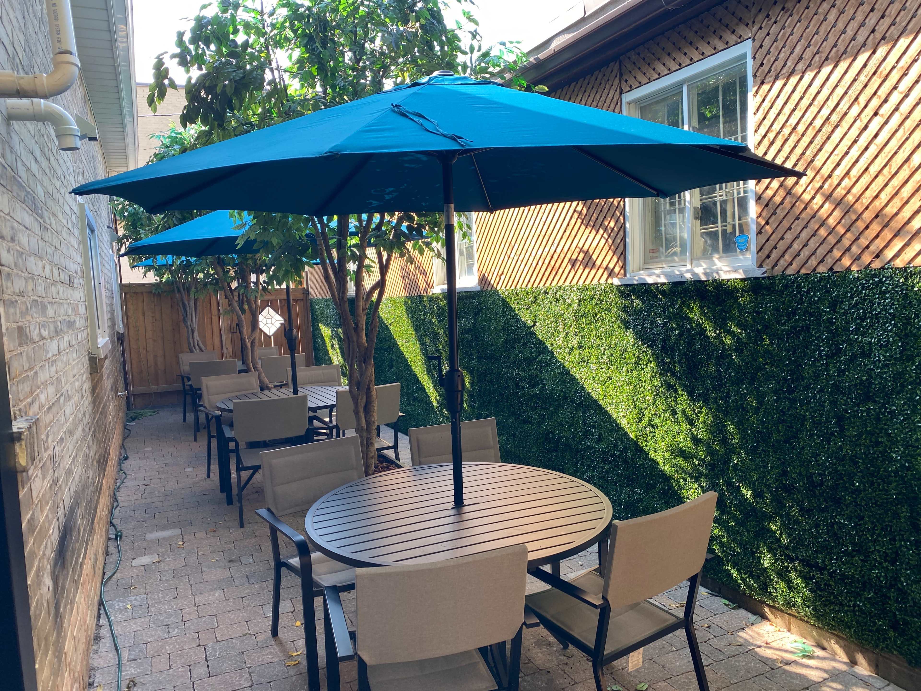 A courtyard features several round tables with beige chairs beneath large blue umbrellas, surrounded by green hedges and brick walls.