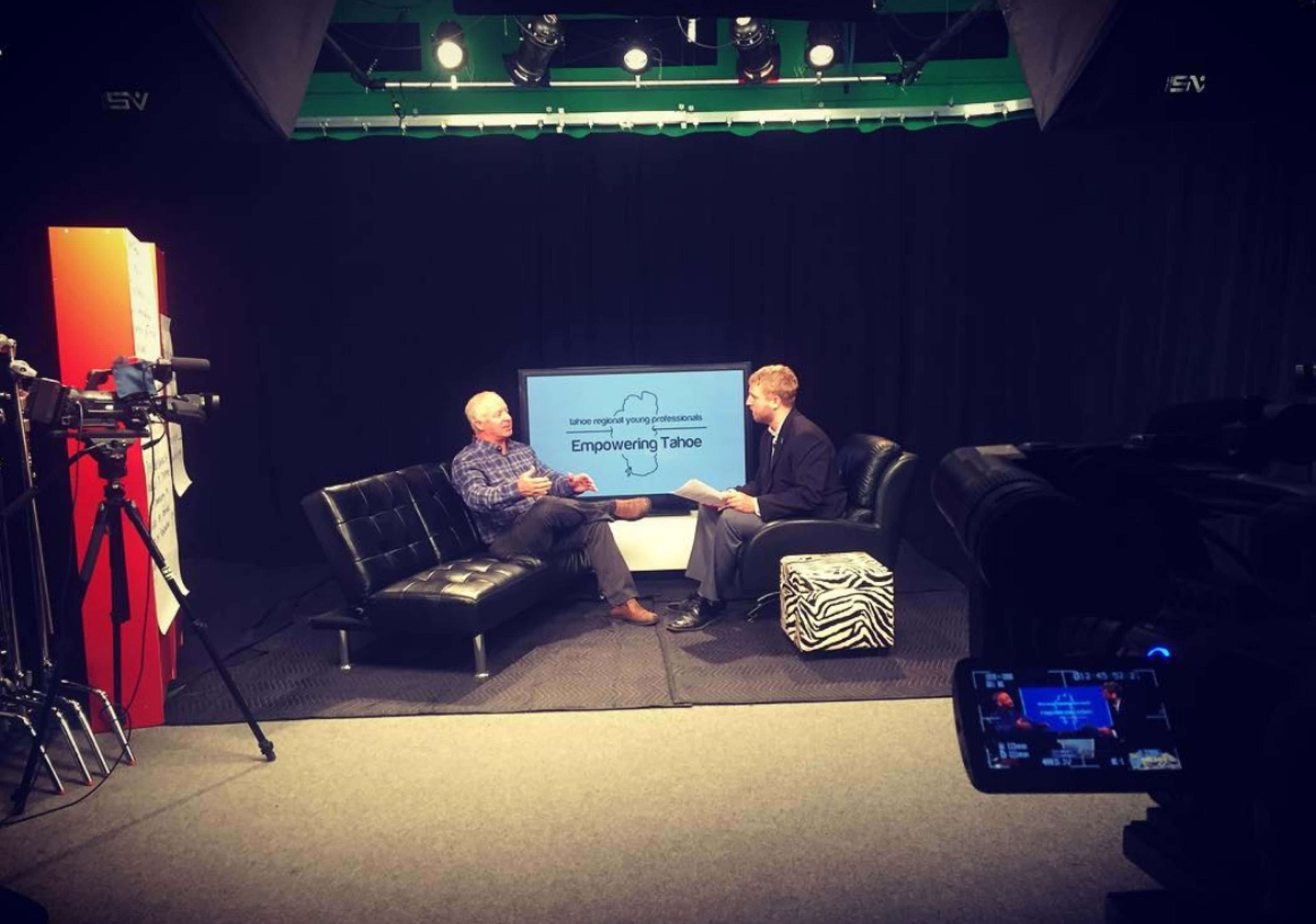 A television studio features two men seated on a couch discussing "Empowering Tahoe," with cameras and lights positioned around them.