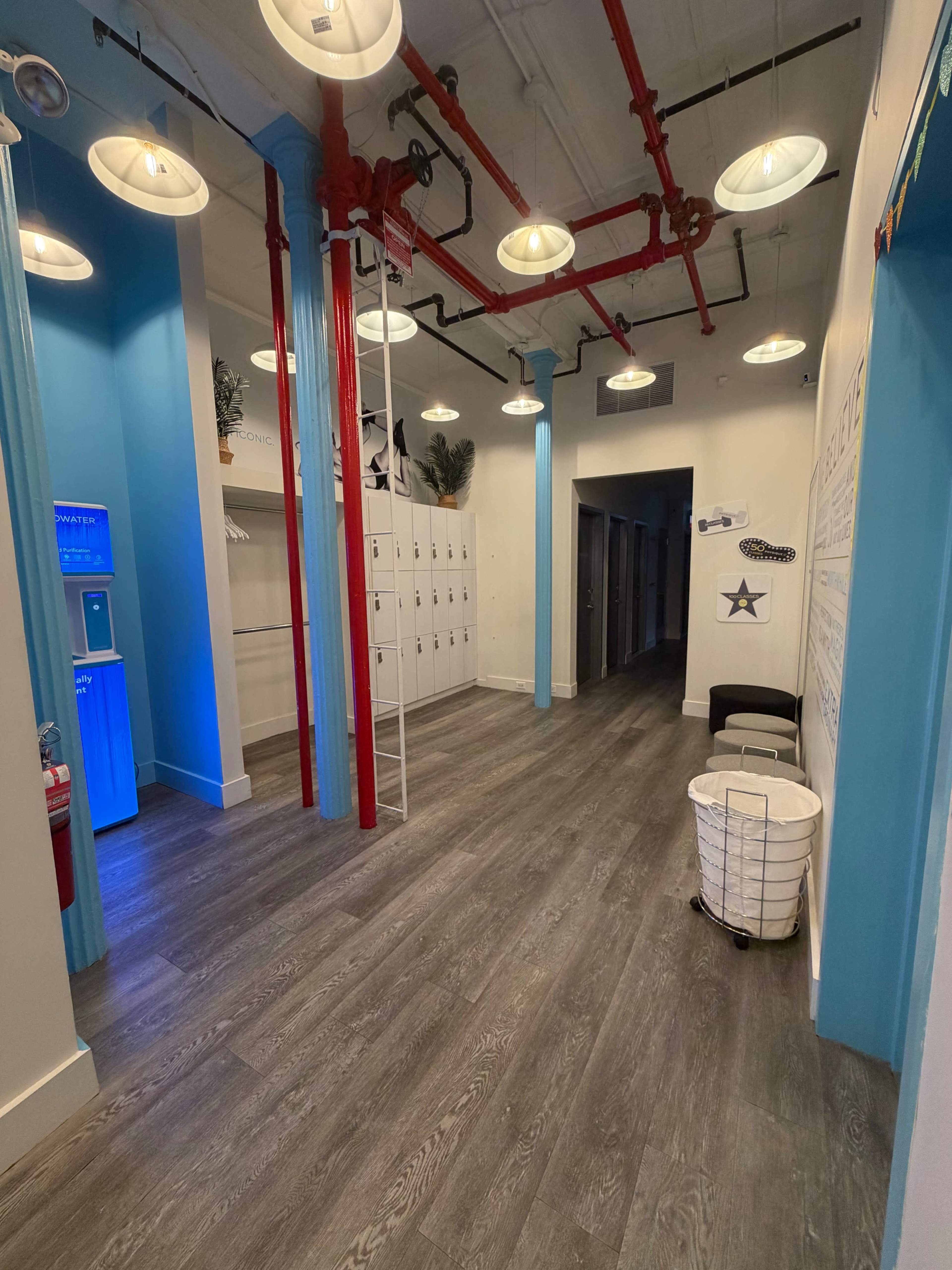 The image shows a clean, well-lit hallway with blue and red accent pillars, lockers on one side, and a water dispenser on the left.