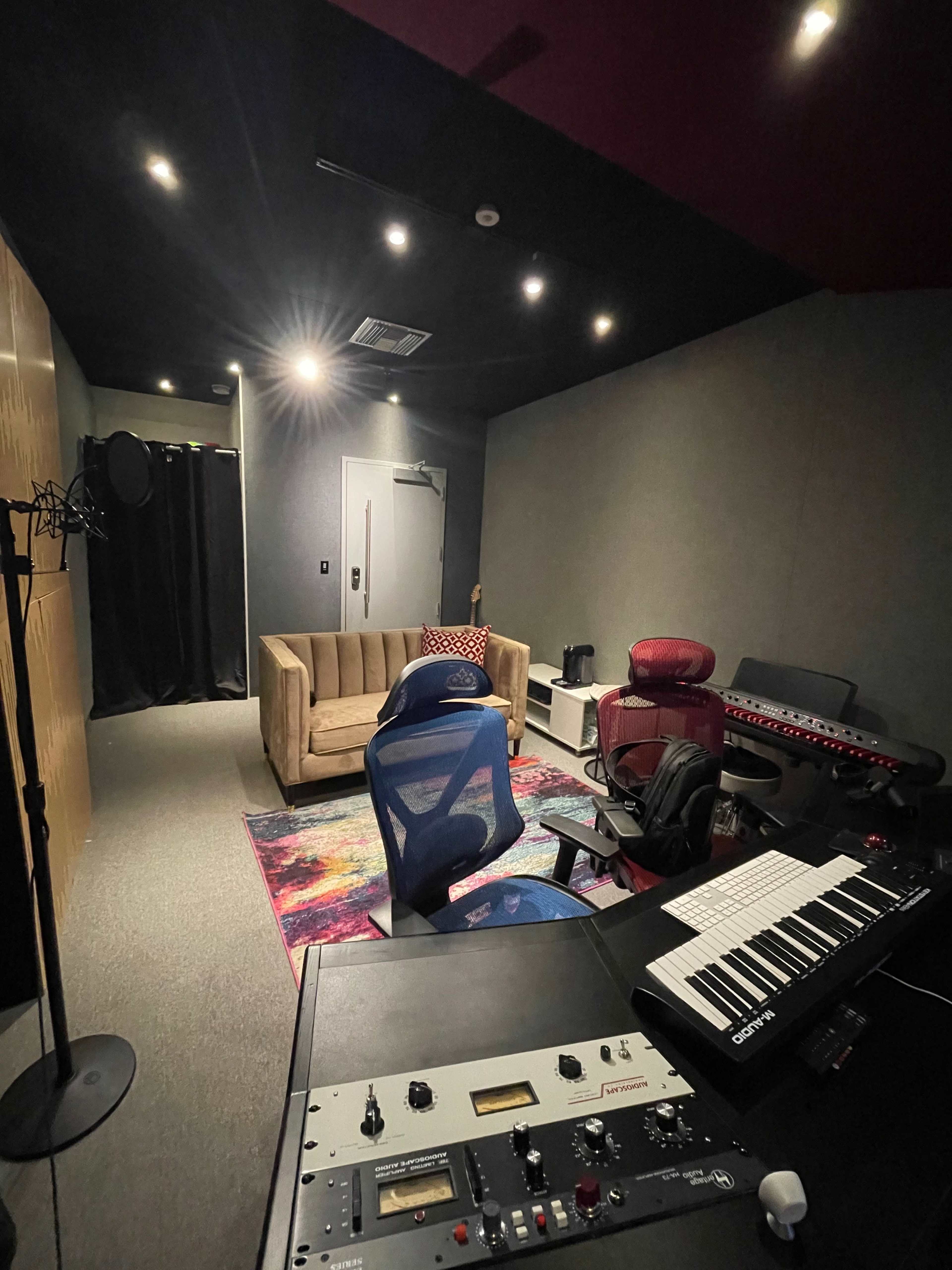 A small recording studio featuring a keyboard setup, a comfortable couch, and several office chairs, with dim lighting and soundproof walls.