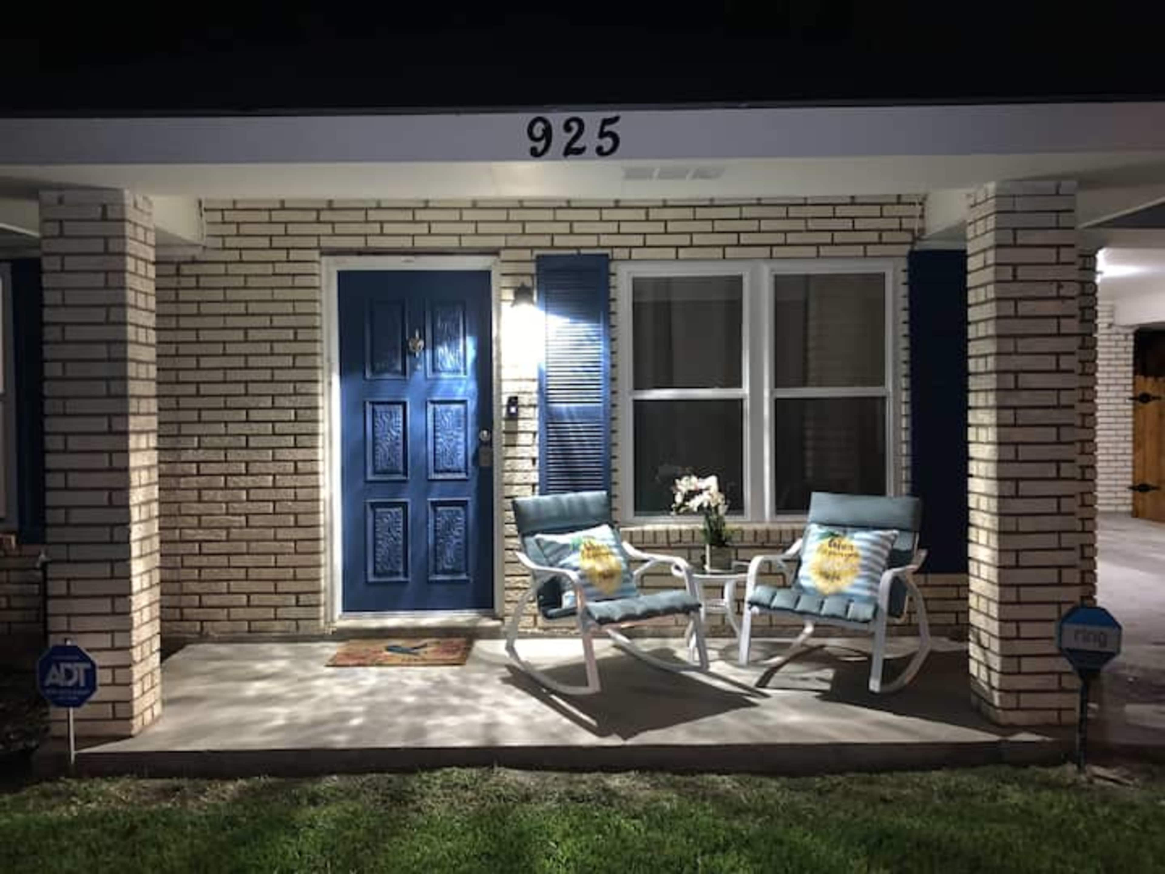 A house with a blue door, two light blue chairs on the porch, and a flower arrangement on a table beside the door.