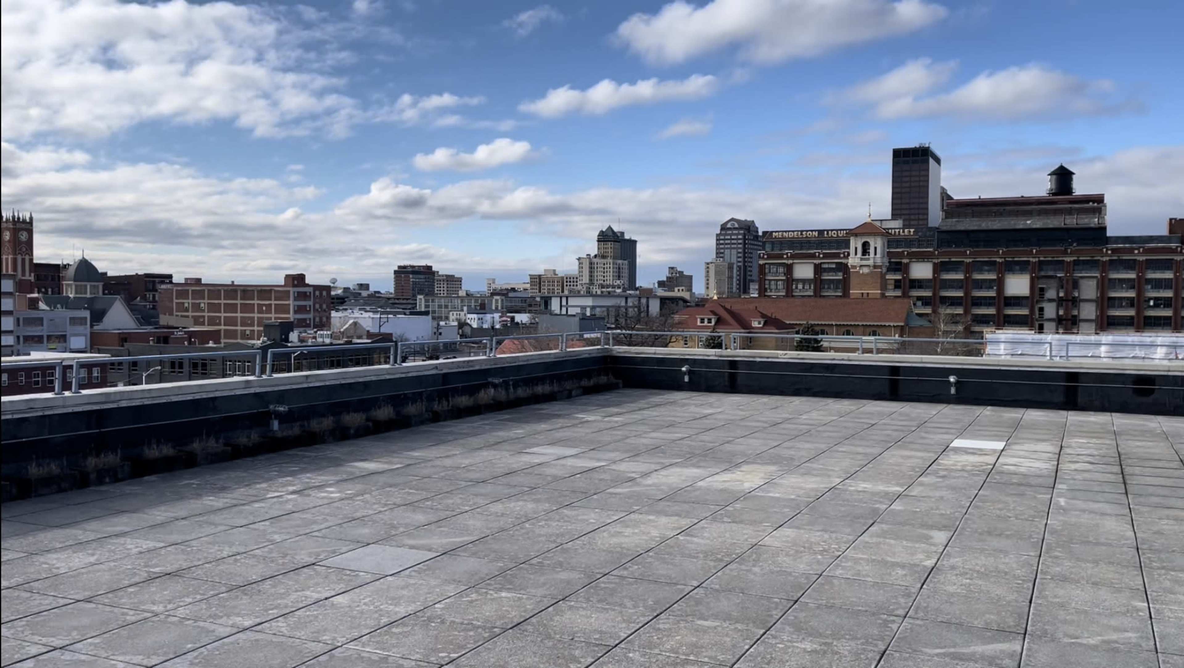 A large, empty rooftop terrace with a concrete surface overlooks a city skyline under a partly cloudy sky.