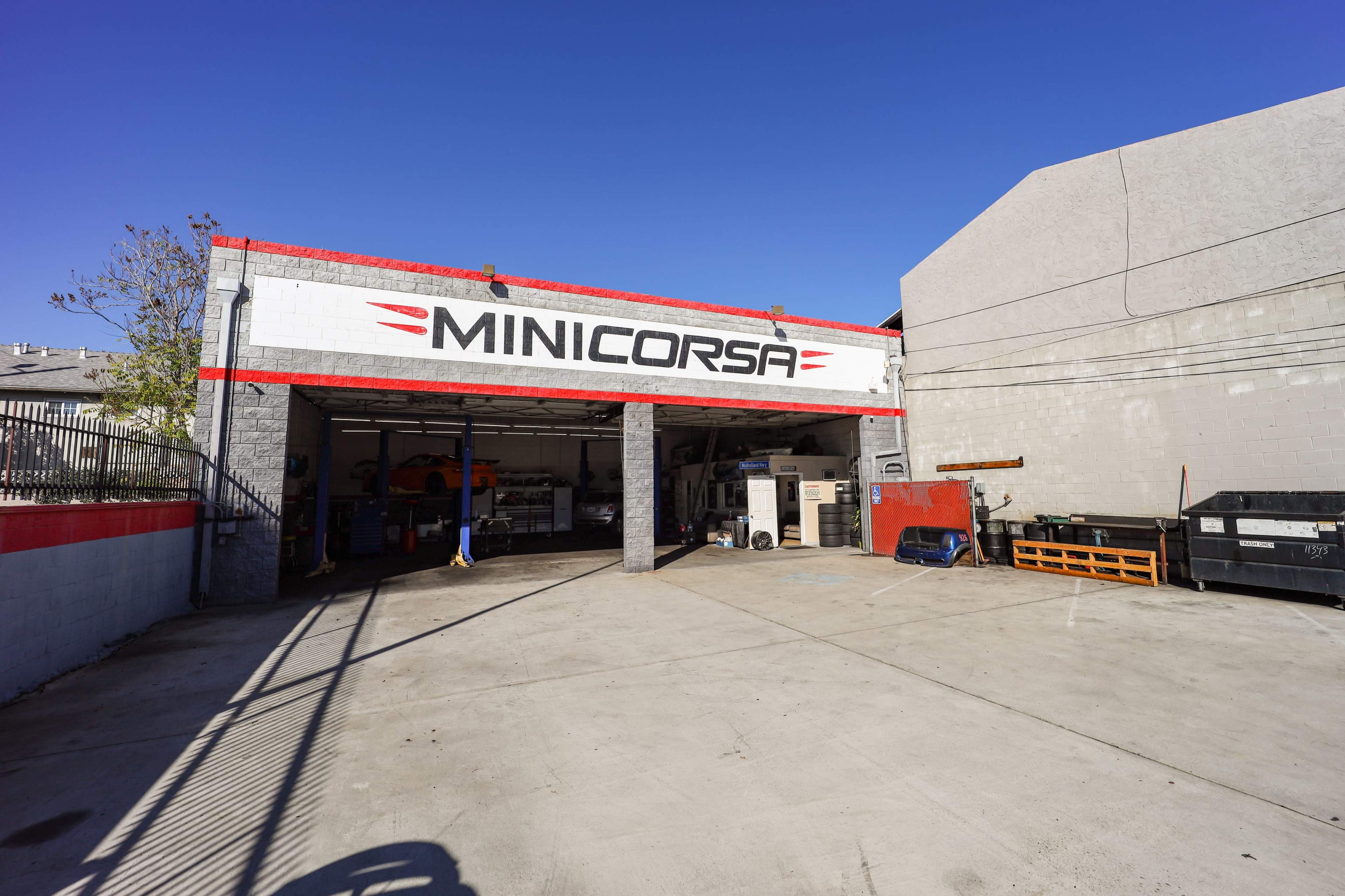 A garage with a sign reading "MINICORSA" above an open entrance, revealing various tools and equipment inside, set against a clear blue sky.