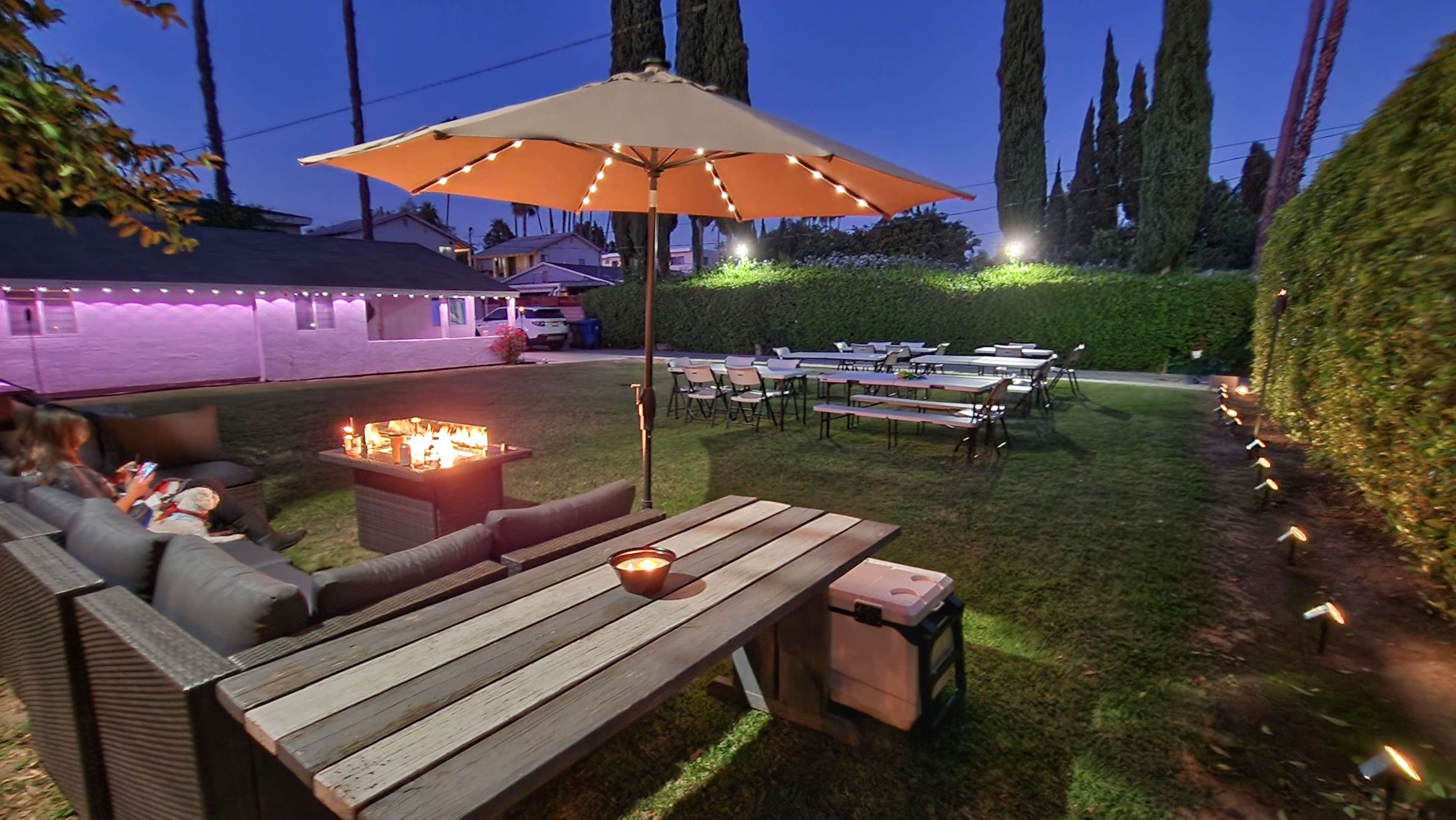 A backyard area features a large table with chairs, a fire pit, and an umbrella, illuminated by string lights and surrounded by greenery.