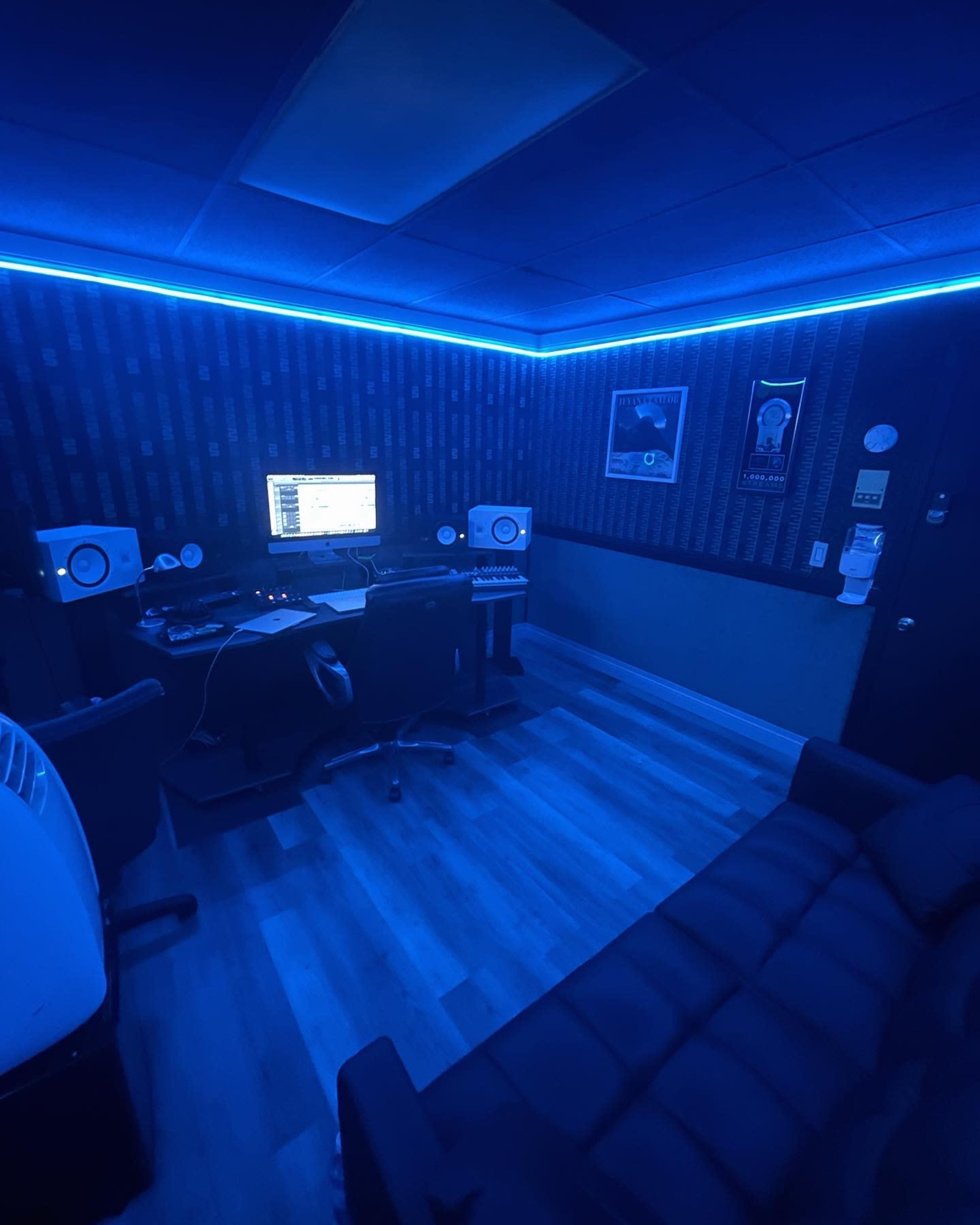A dimly lit music production studio with blue ambient lighting, featuring a computer set up with speakers and a black couch.