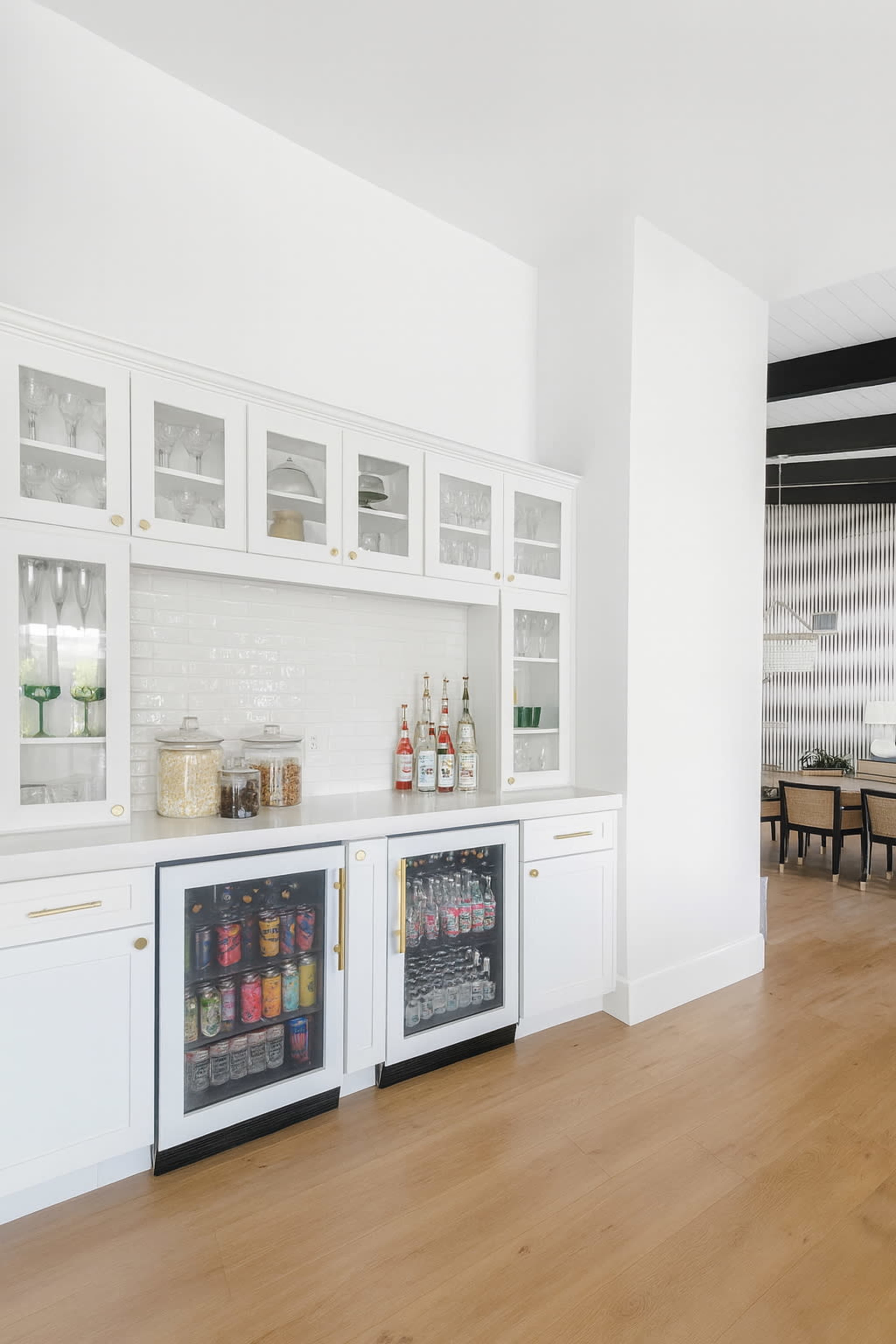 A modern kitchen features a white cabinetry unit with glass doors, showcasing glassware and decorative items, and two under-counter refrigerators displaying various beverages.
