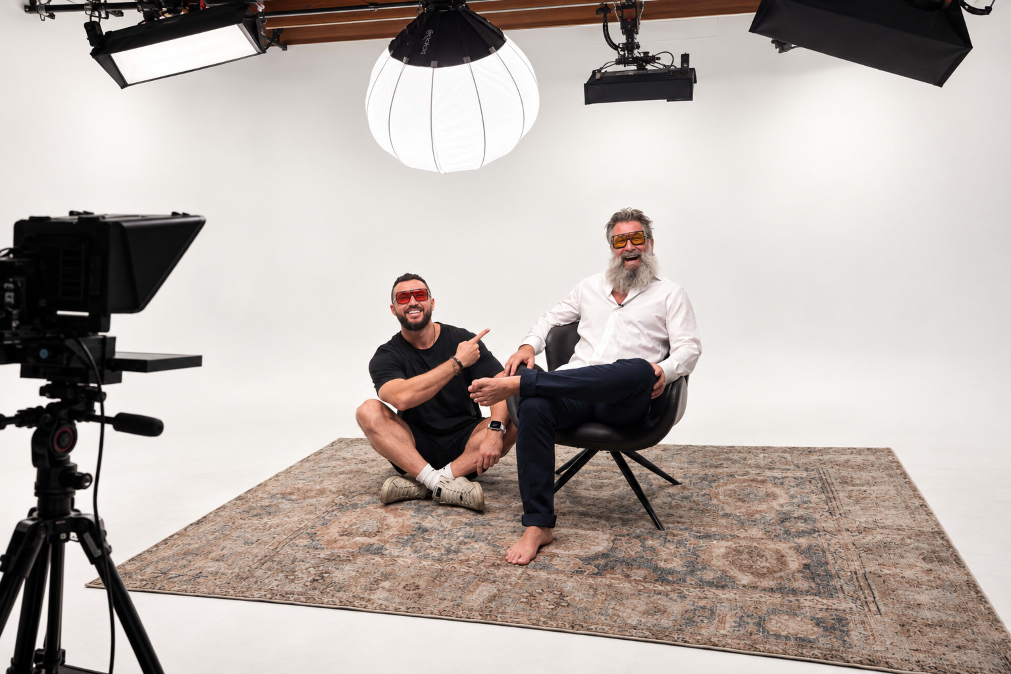 Two men sit in a studio setting on a patterned rug, with one man in a chair and the other seated on the floor, both wearing sunglasses and appearing relaxed.