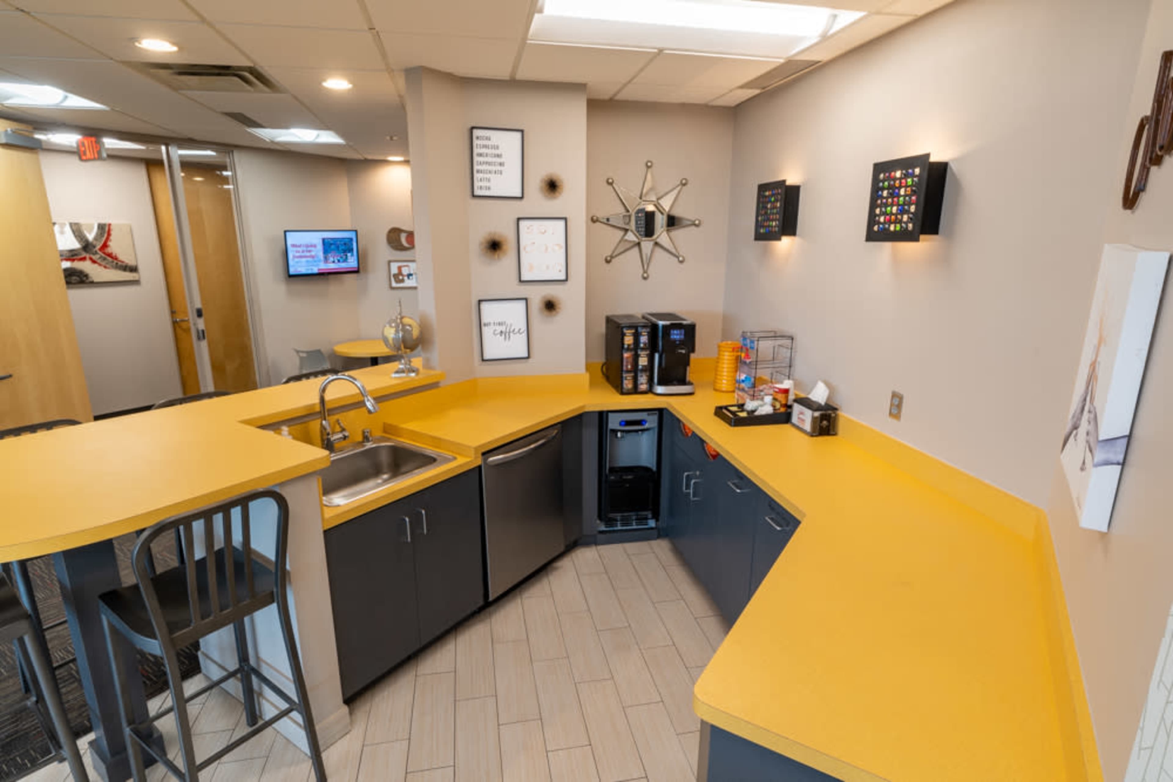 A modern kitchenette area with yellow countertops, a sink, a coffee station, and wall-mounted decor.
