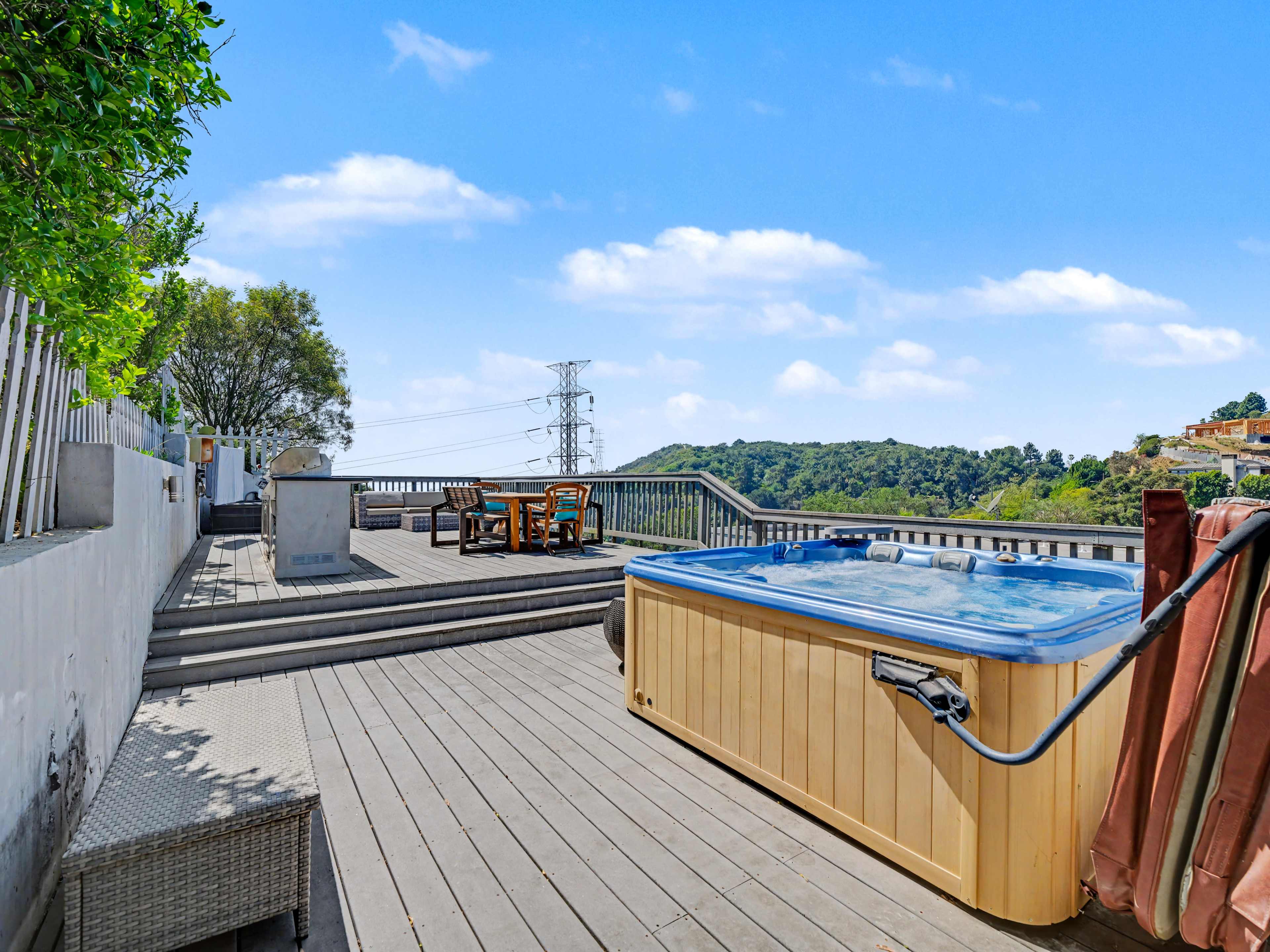 A wooden deck features a hot tub and outdoor seating area, overlooking a hillside and power lines in the background.