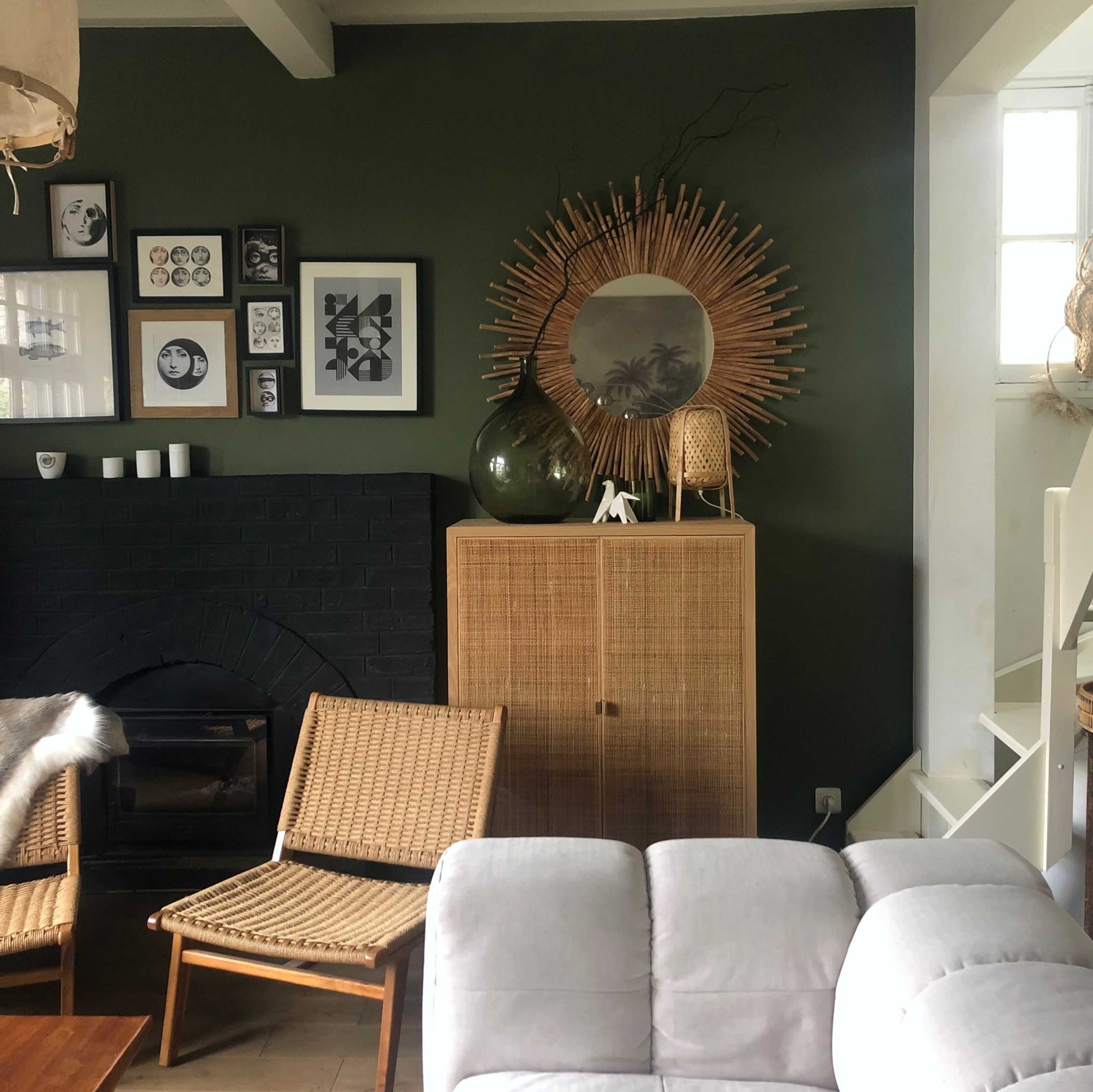 The scene features a living room with a dark green wall, framed artwork, a round mirror, a rattan cabinet, and seating arrangements with a fireplace.