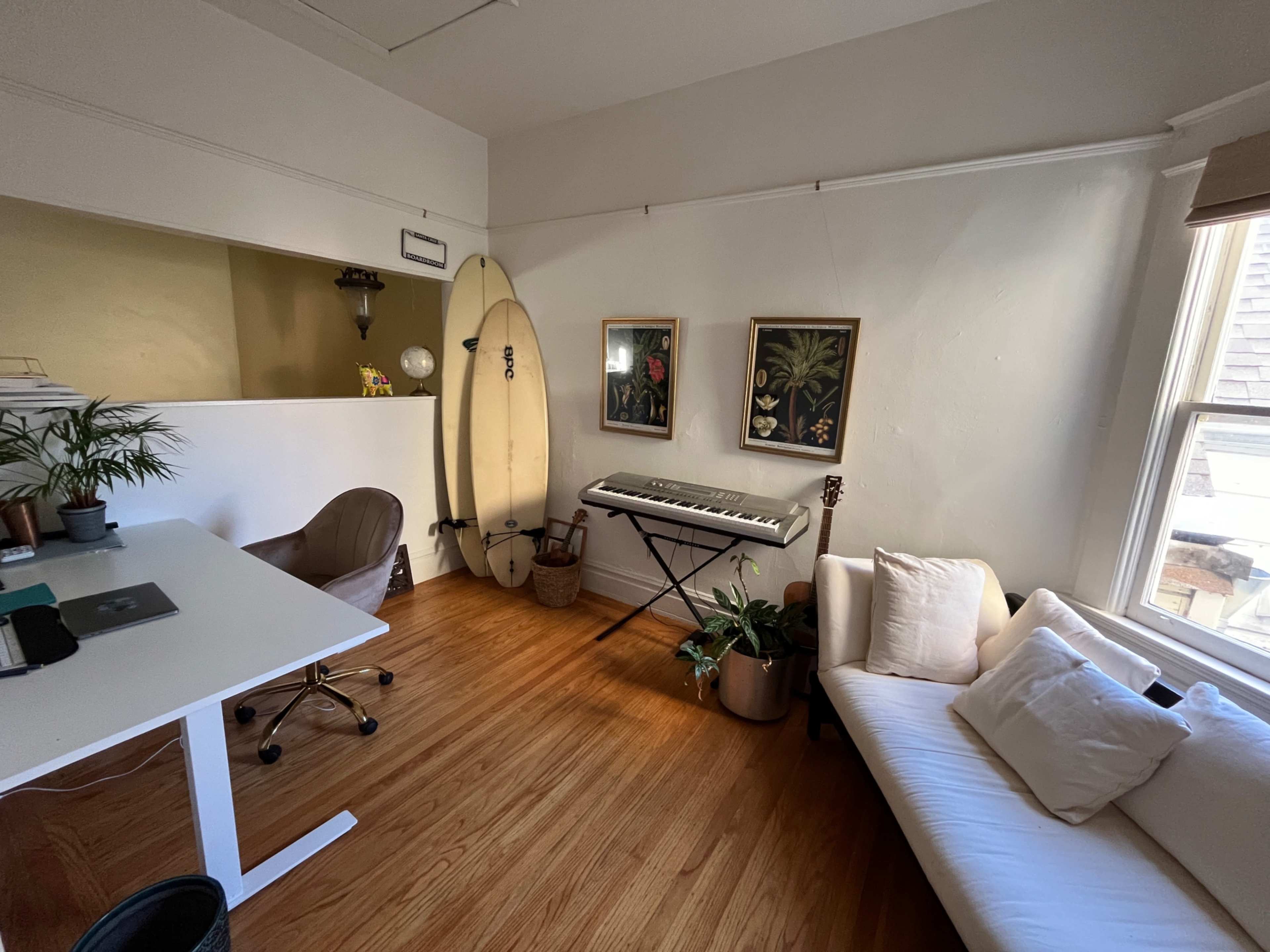 The room features a white desk, a surfboard, a keyboard, framed artwork, and a sofa, with wooden flooring and natural light coming through the windows.