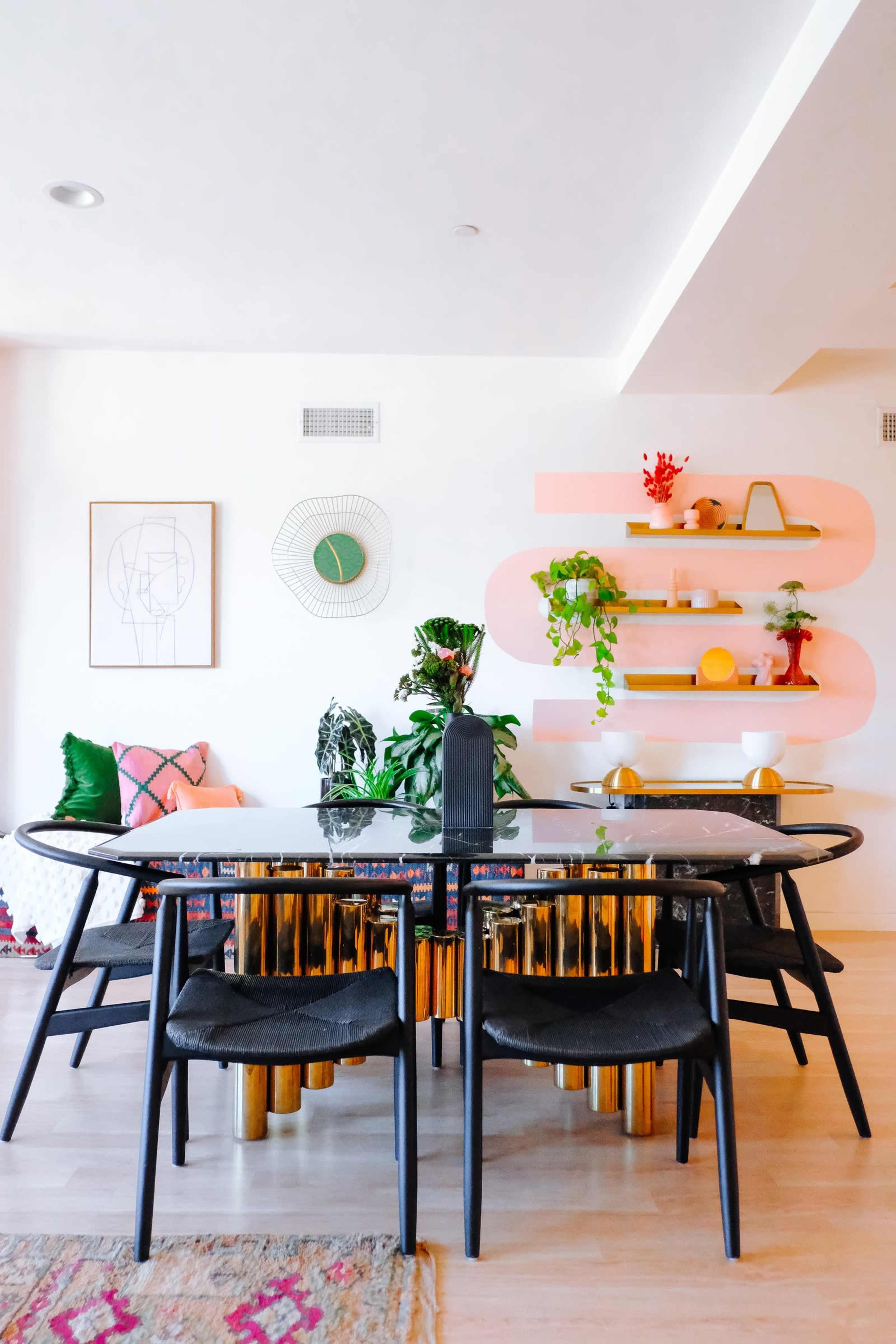 A modern dining area features a round table surrounded by black chairs, with plants and decorative items displayed on pink shelves in the background.