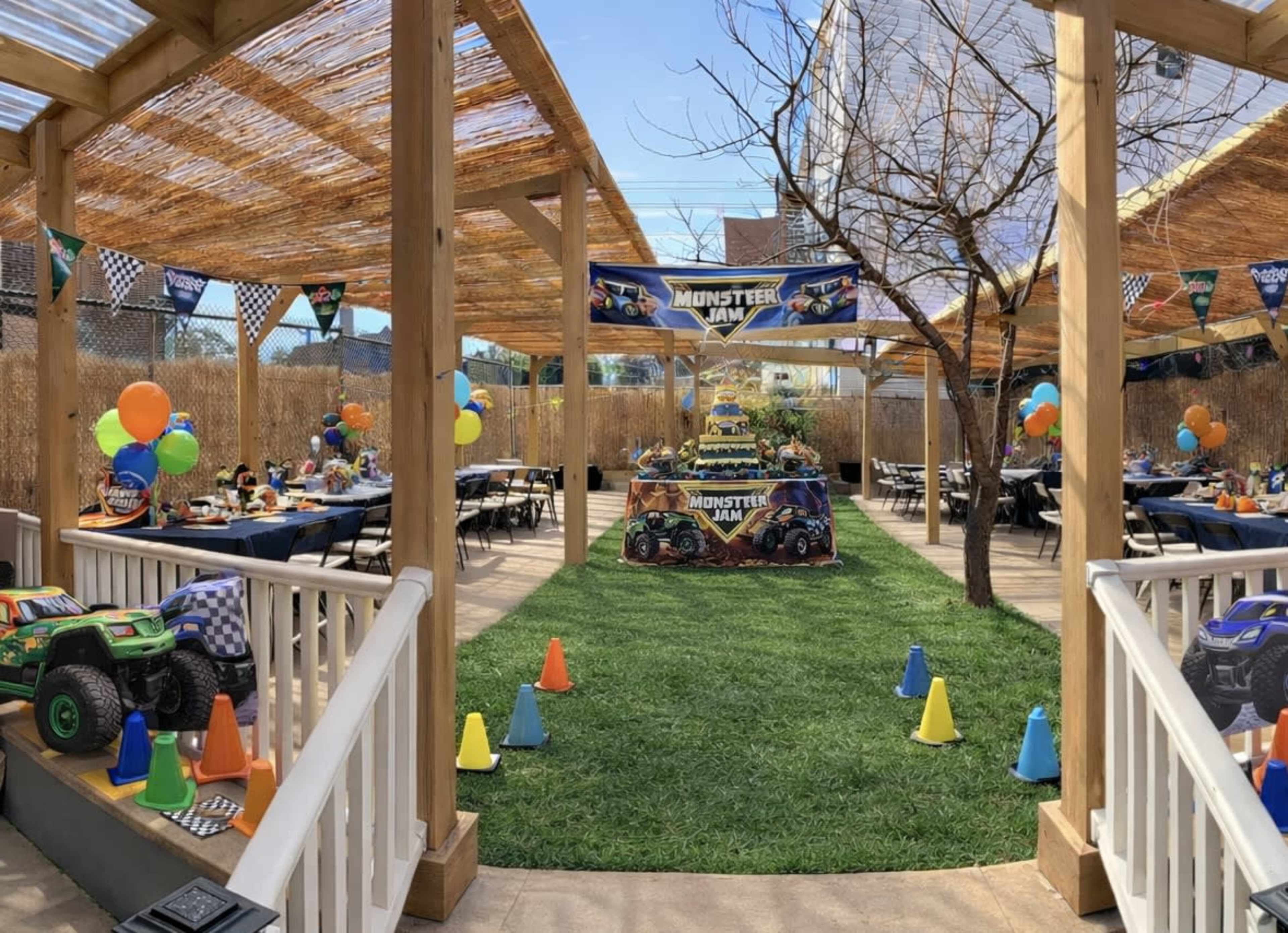 The scene features a decorated outdoor area set up for a party, with tables arranged around a grassy space, colorful balloons, and a monster truck-themed display.