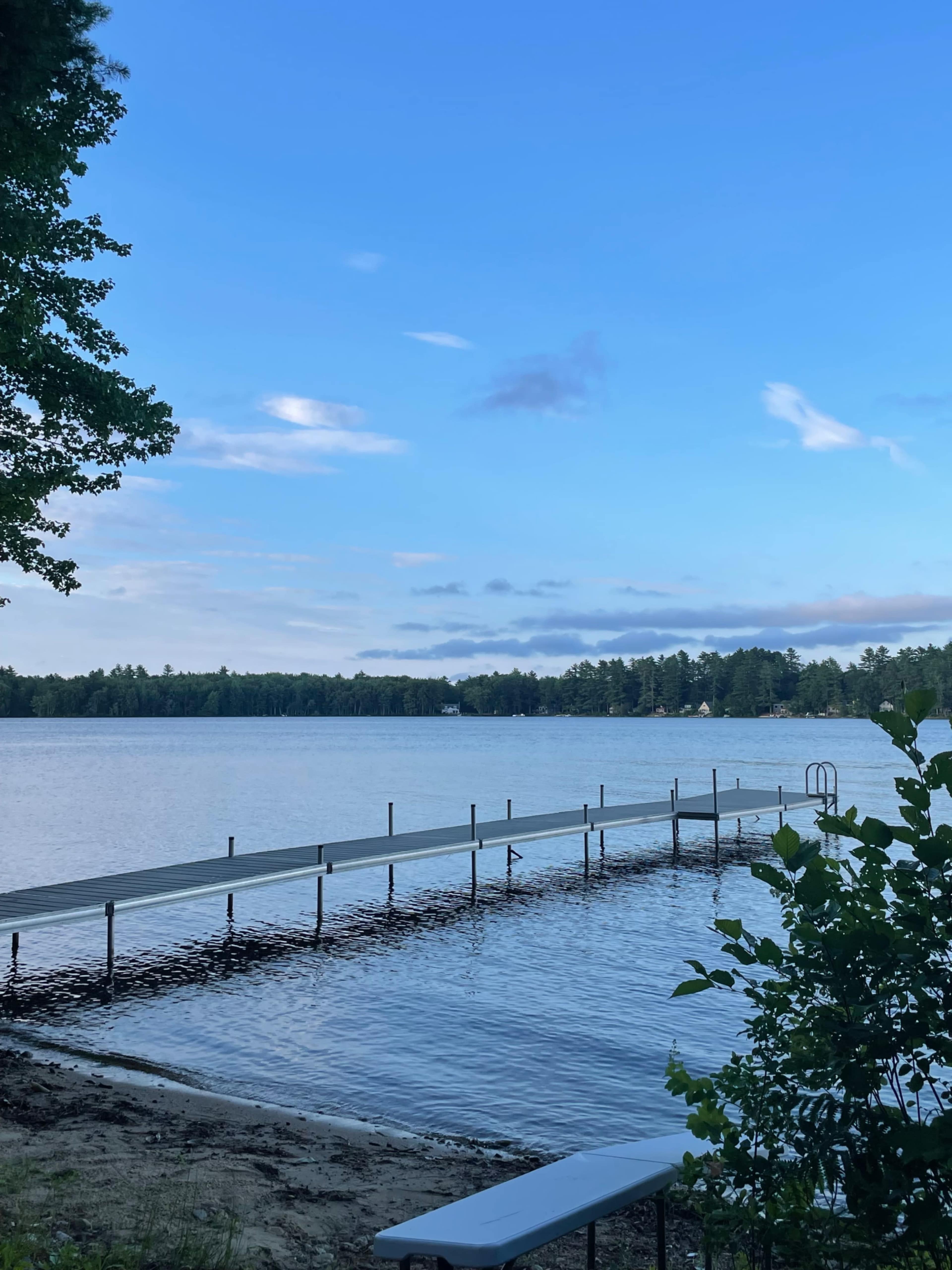 A wooden dock extends into a calm lake surrounded by trees under a blue sky with scattered clouds.