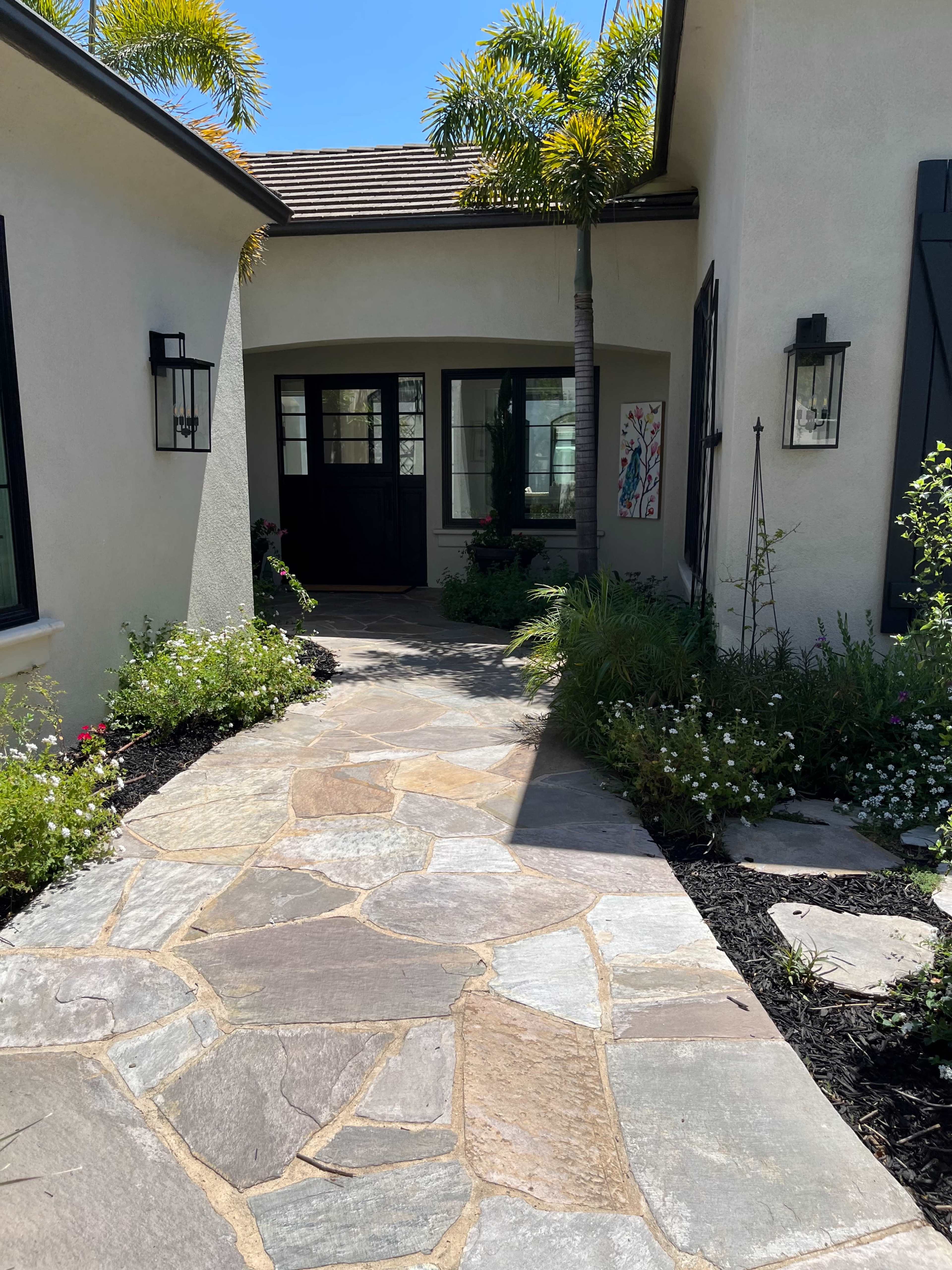 A stone pathway leads to a double-door entrance surrounded by landscaped gardens and potted plants.