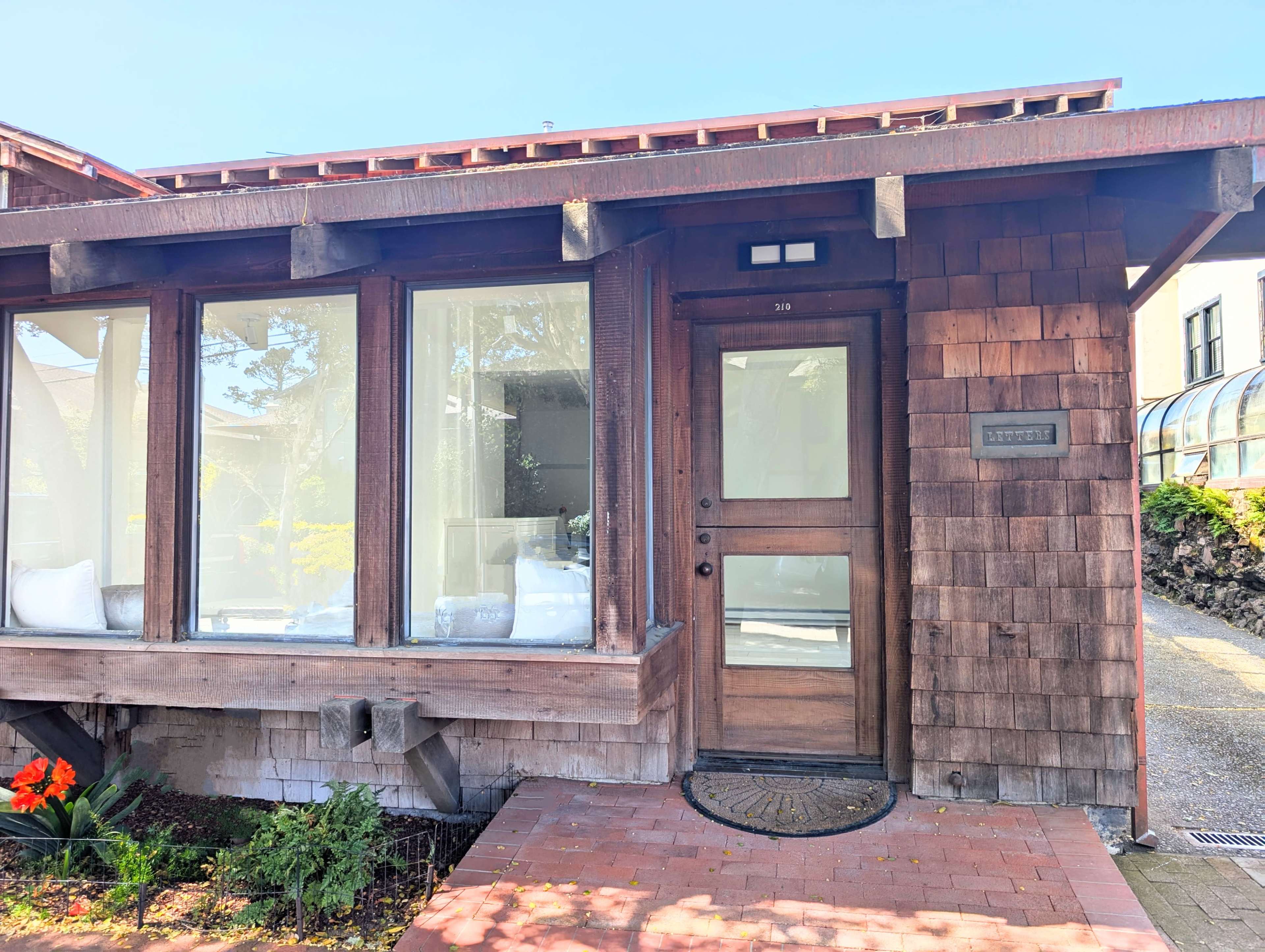 The image shows the entrance of a wooden house with large windows and a brick pathway leading to the front door.
