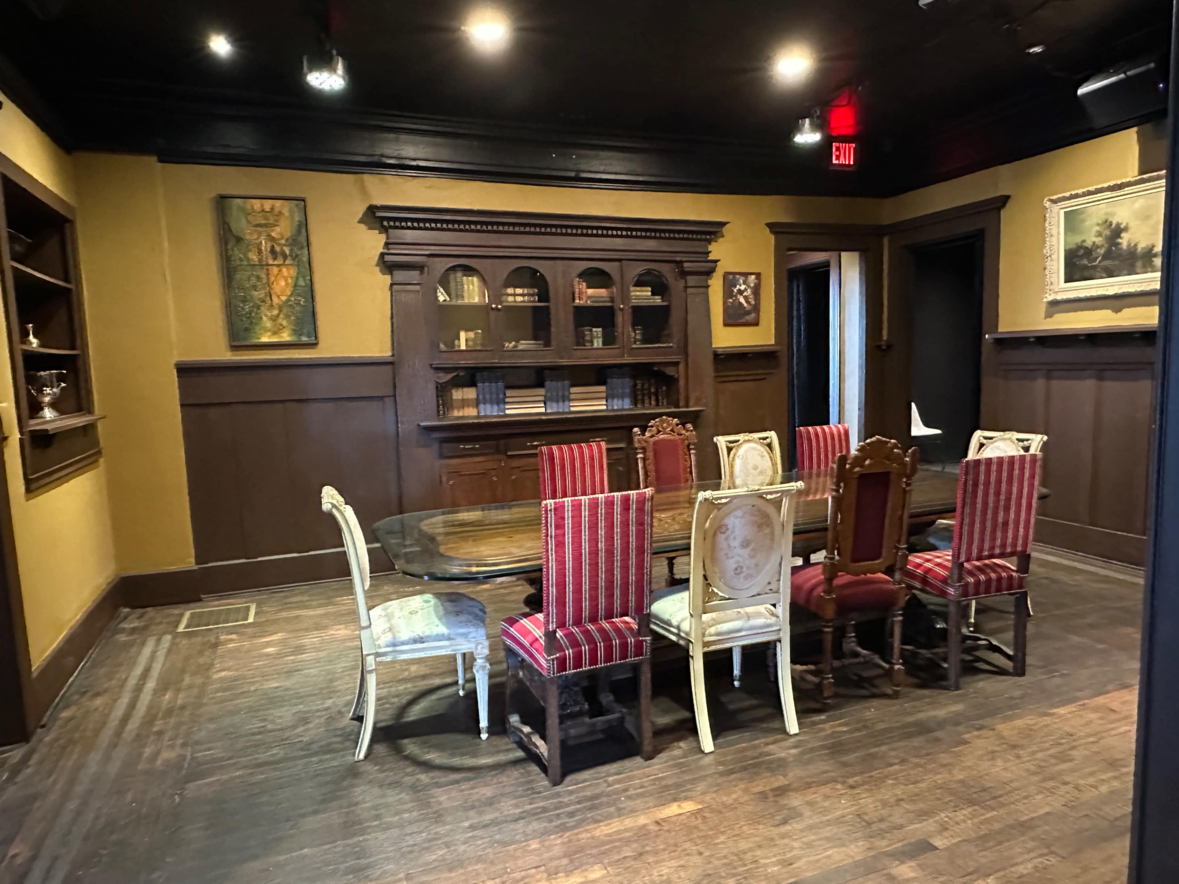 The image shows a dining room with a large wooden table surrounded by eight ornate chairs, set against a backdrop of dark walls and shelves filled with books and decorative items.