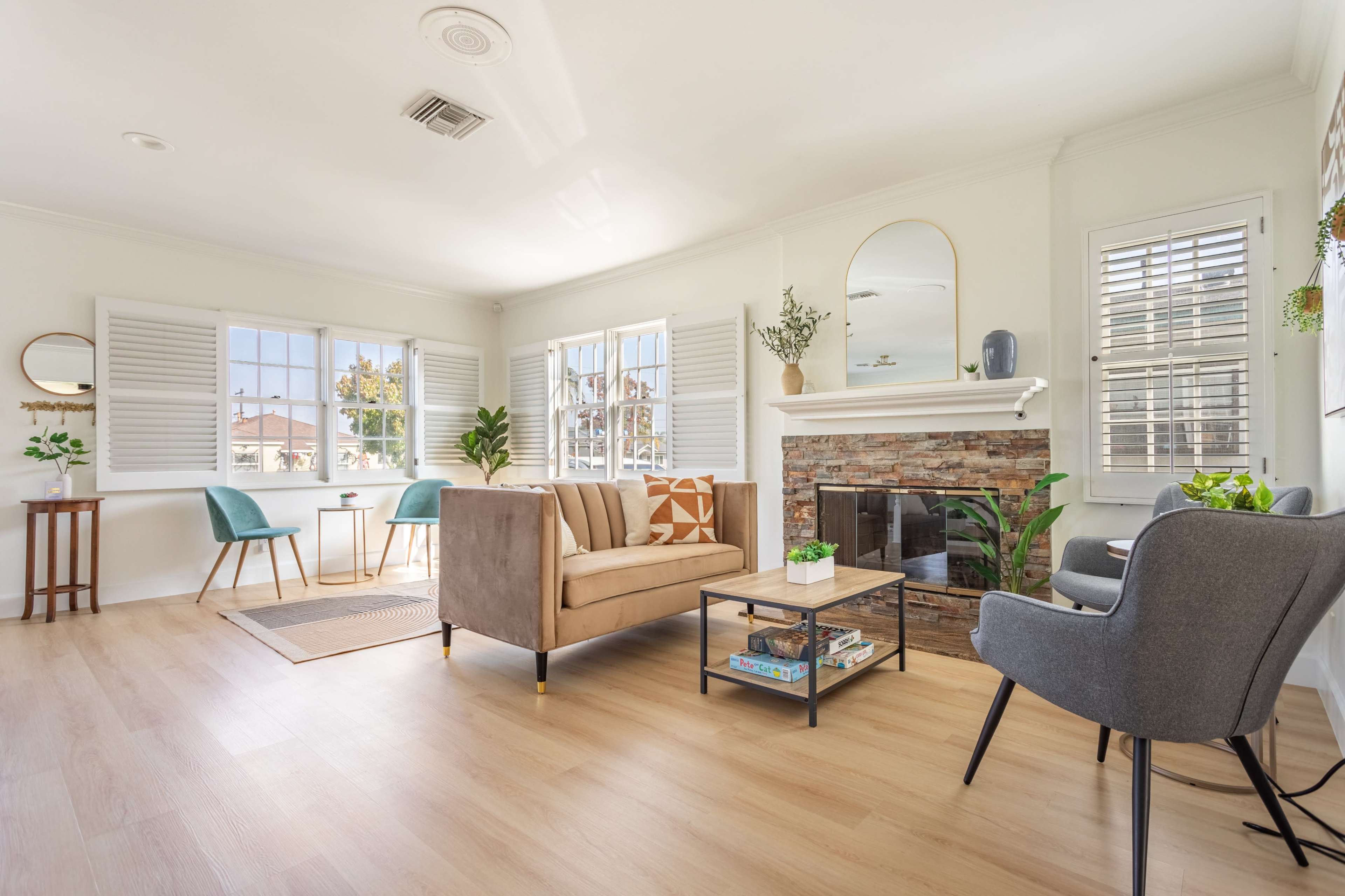 The image shows a light-filled living room featuring a beige sofa, a stone fireplace, two chairs, and large windows with shutters.