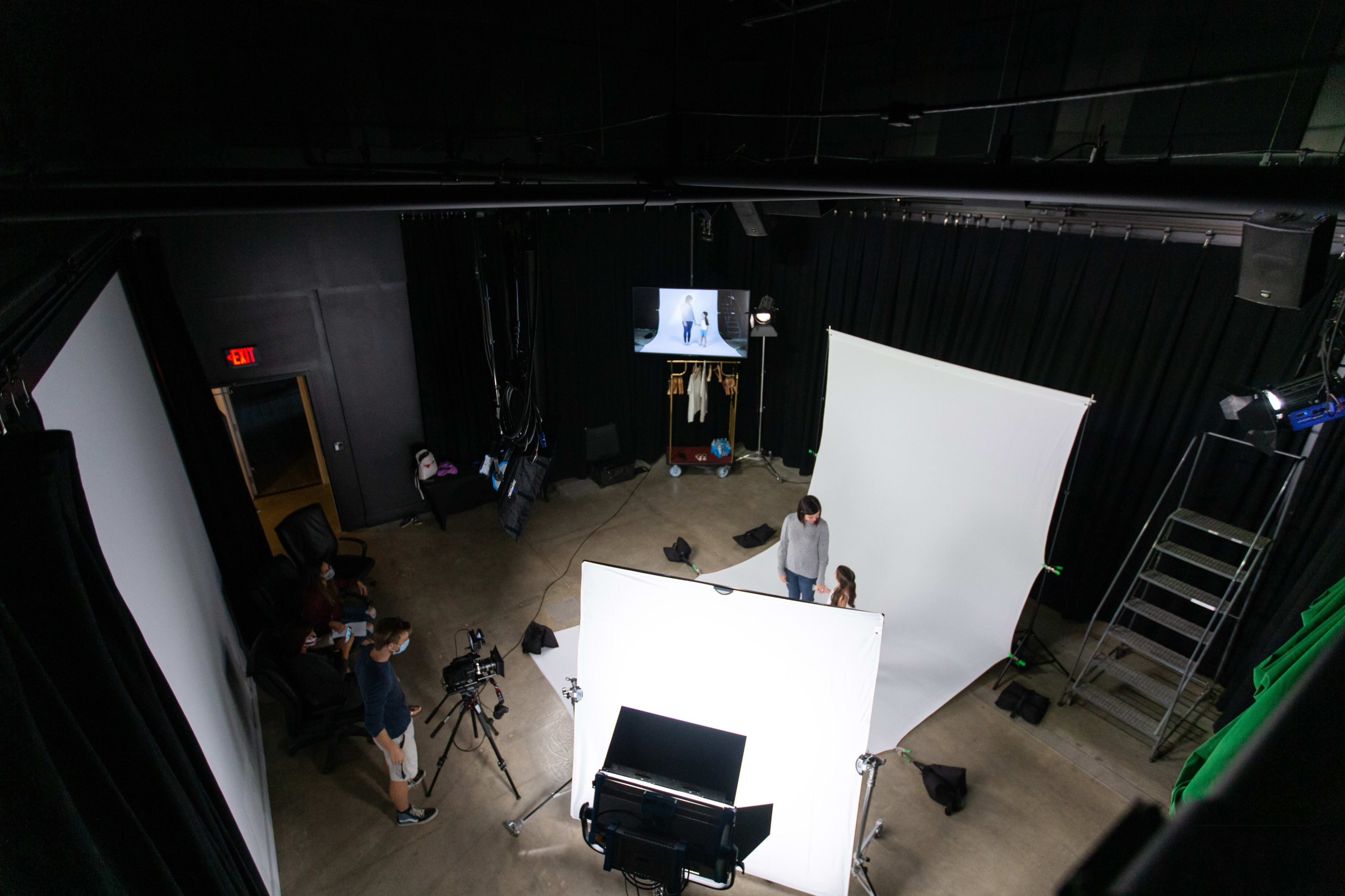A film studio setup features white backdrop screens, cameras, and crew members preparing for a shoot.