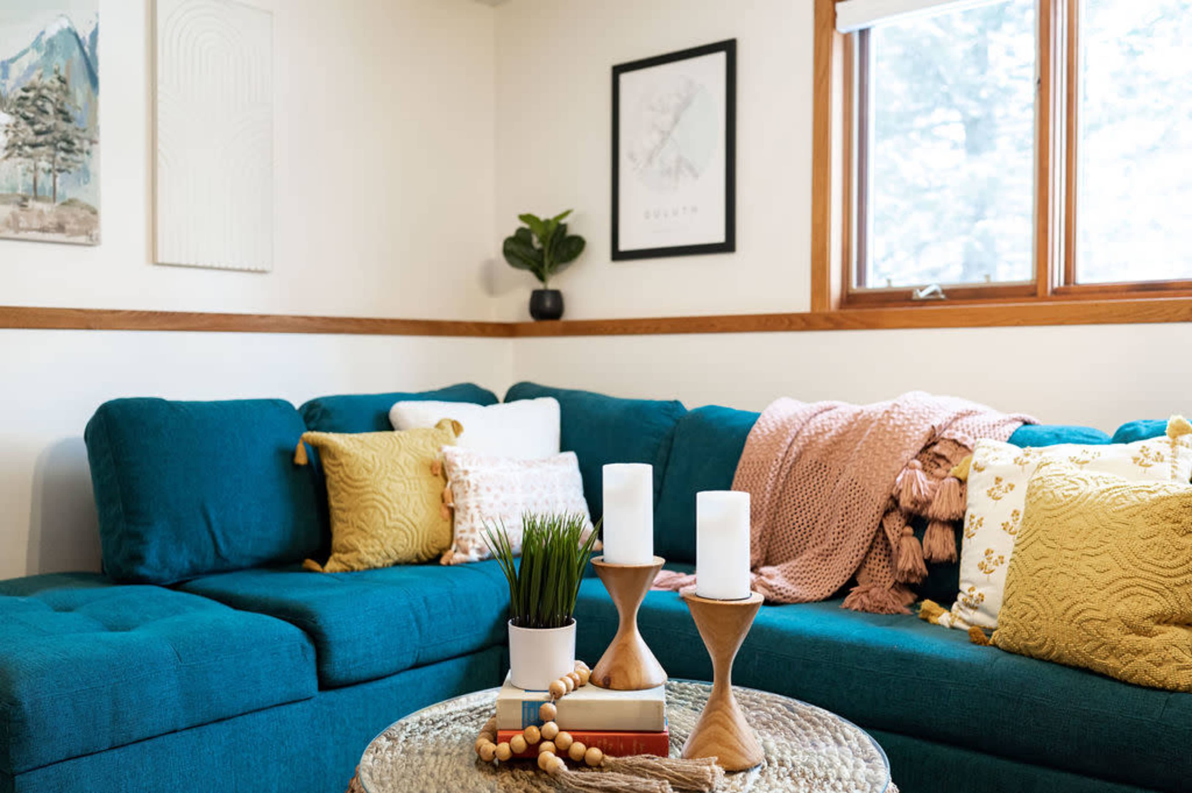 A cozy living room features a teal sectional sofa adorned with colorful pillows, a round coffee table with decorative elements, and a window letting in natural light.