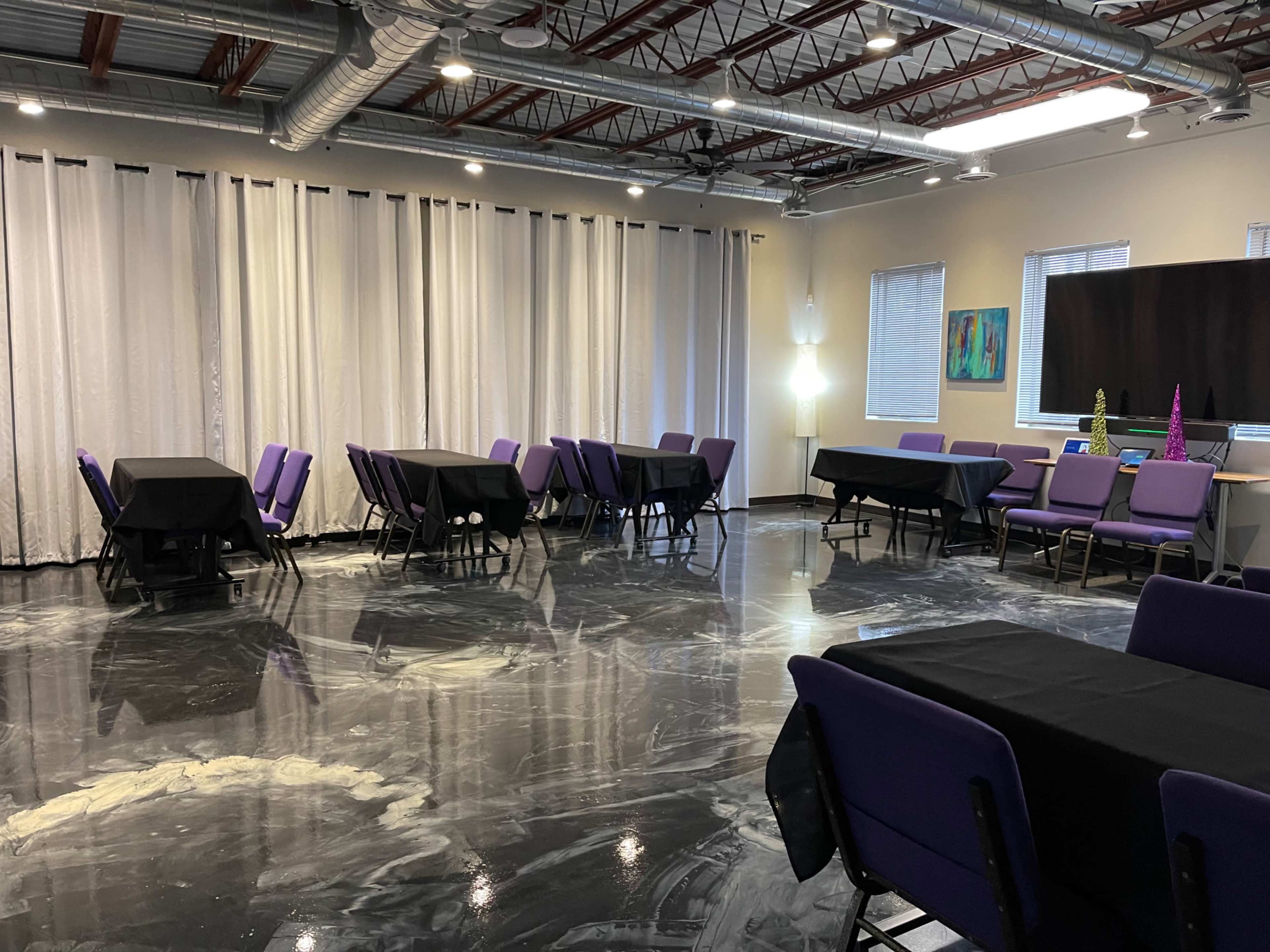 A spacious room set up with purple-covered tables and chairs, with black tablecloths across the tables and large white curtains covering the windows.