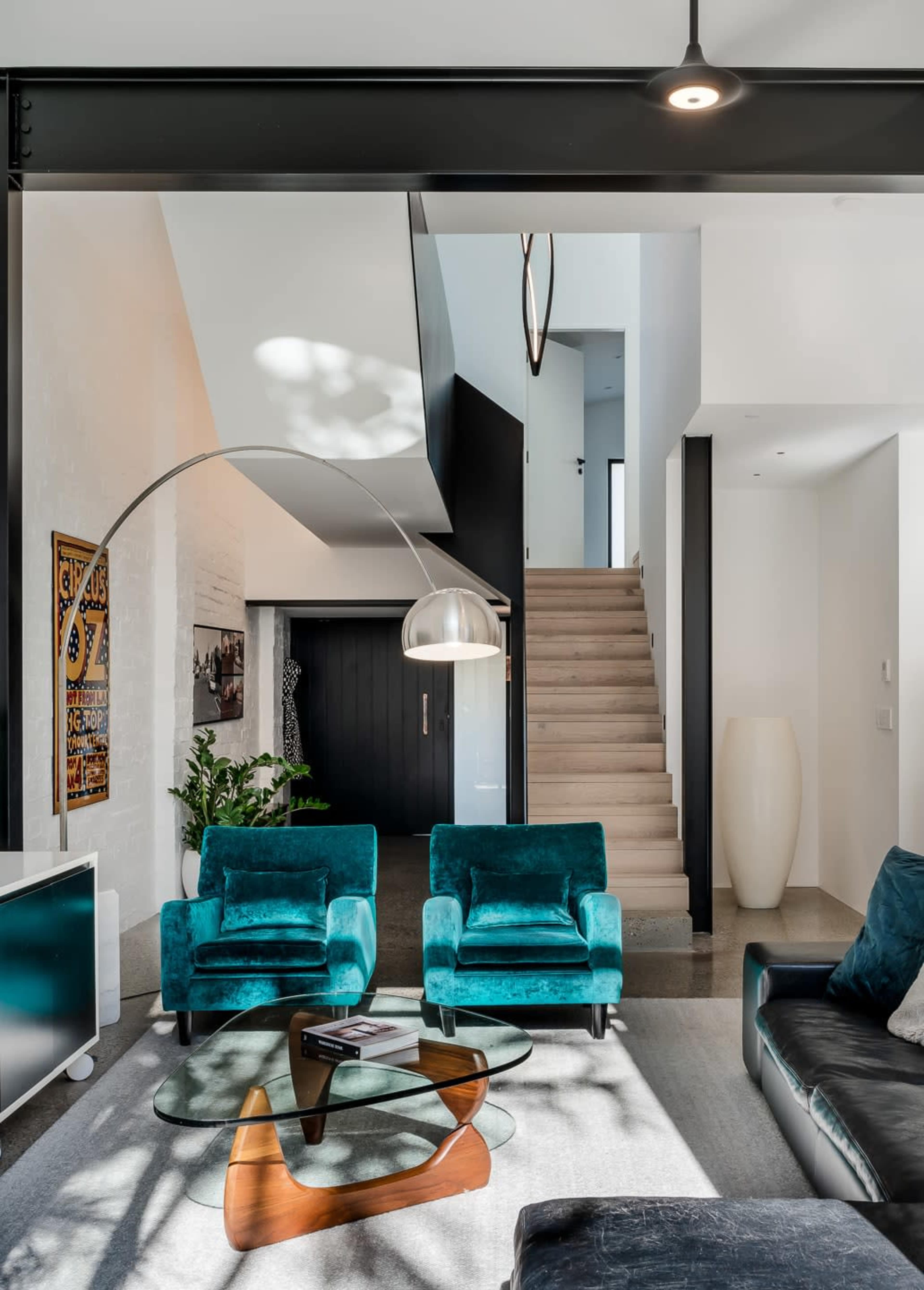 A modern living room featuring two teal armchairs, a glass coffee table, and a staircase leading to an upper level.
