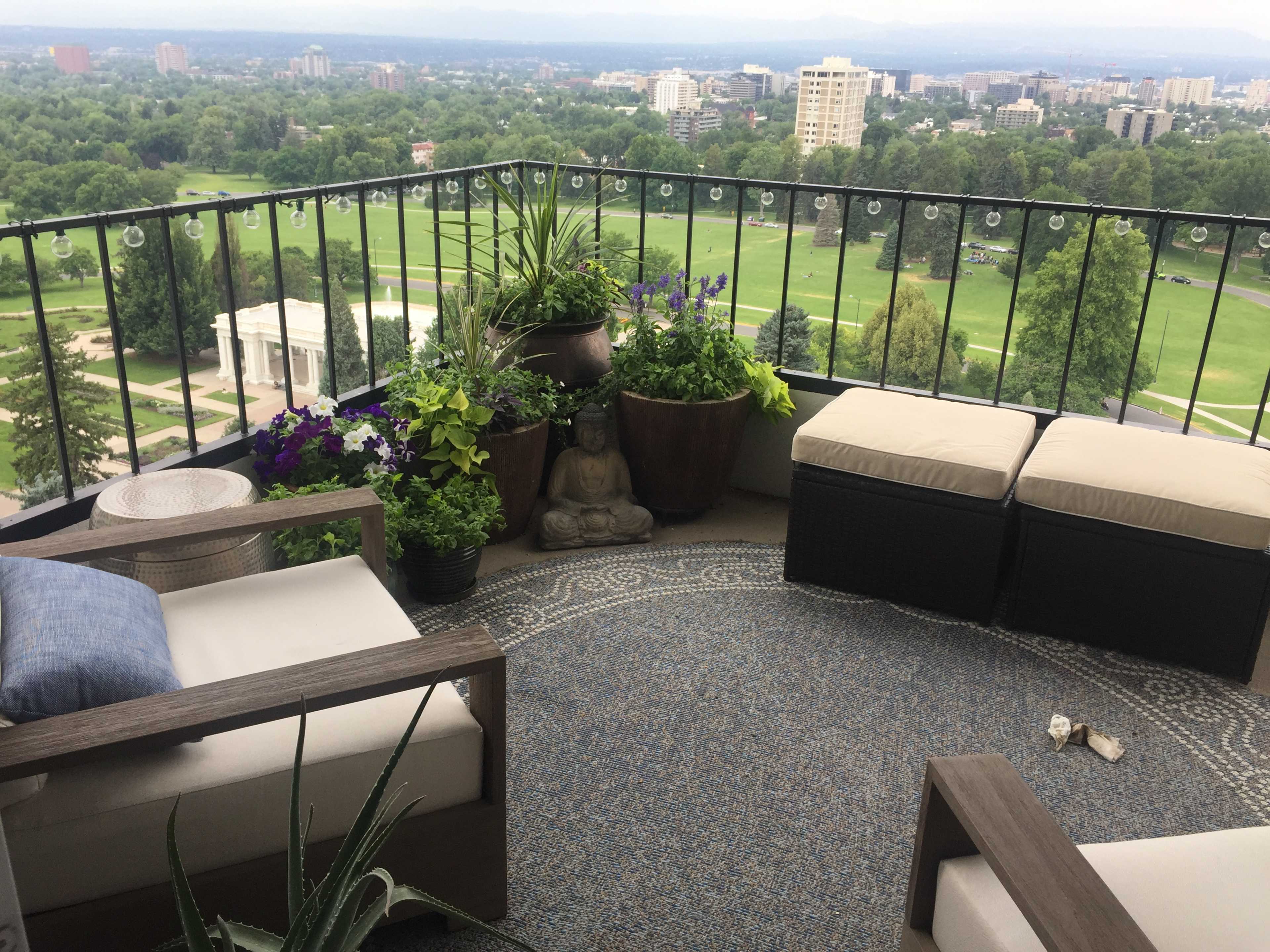 A balcony with potted plants, a small statue, and two seated areas overlooking a green landscape and city skyline.