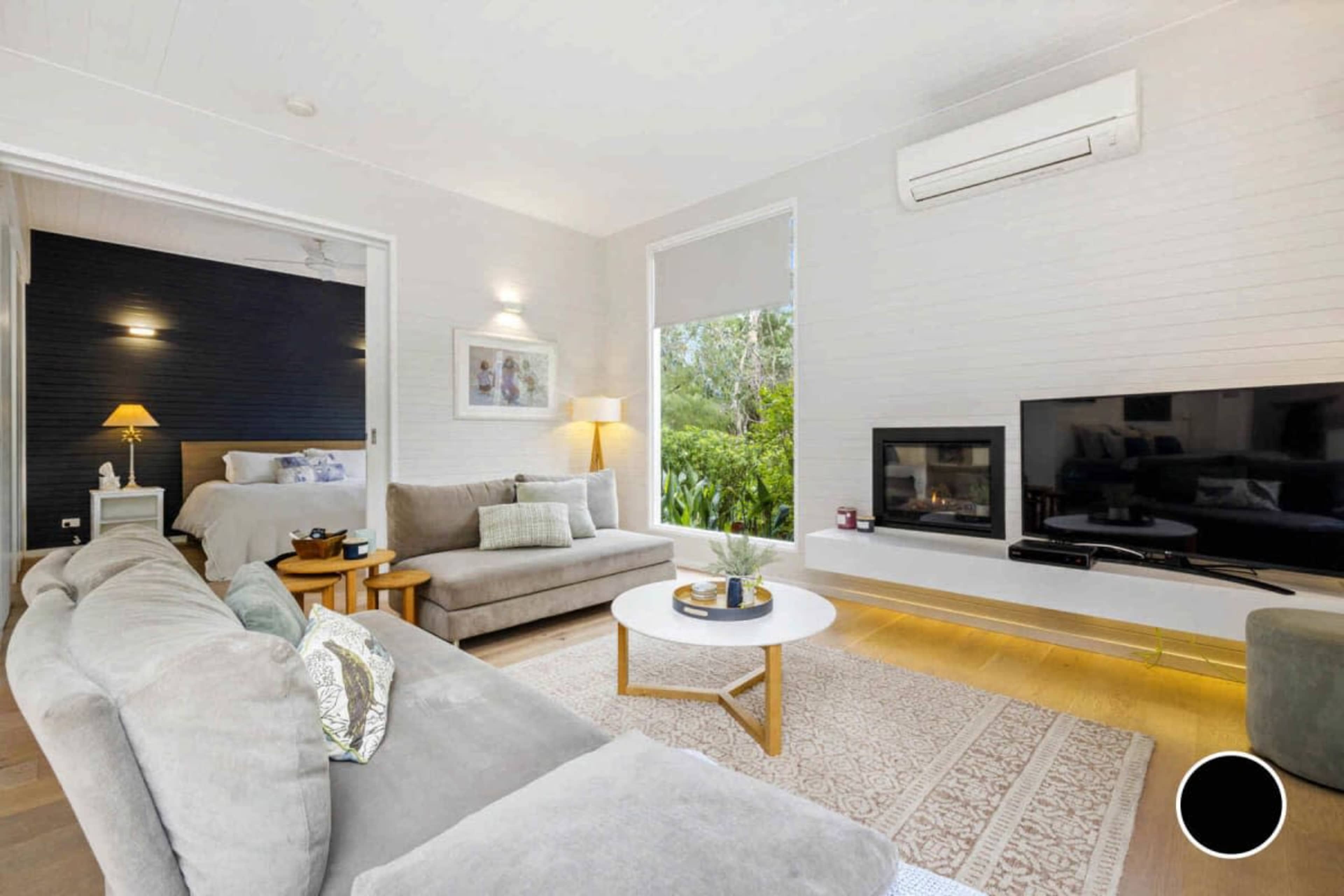 A modern living room with a gray sofa, a circular coffee table, a flat-screen TV, and a view of a bedroom area through an open doorway, featuring a bed with a lamp.