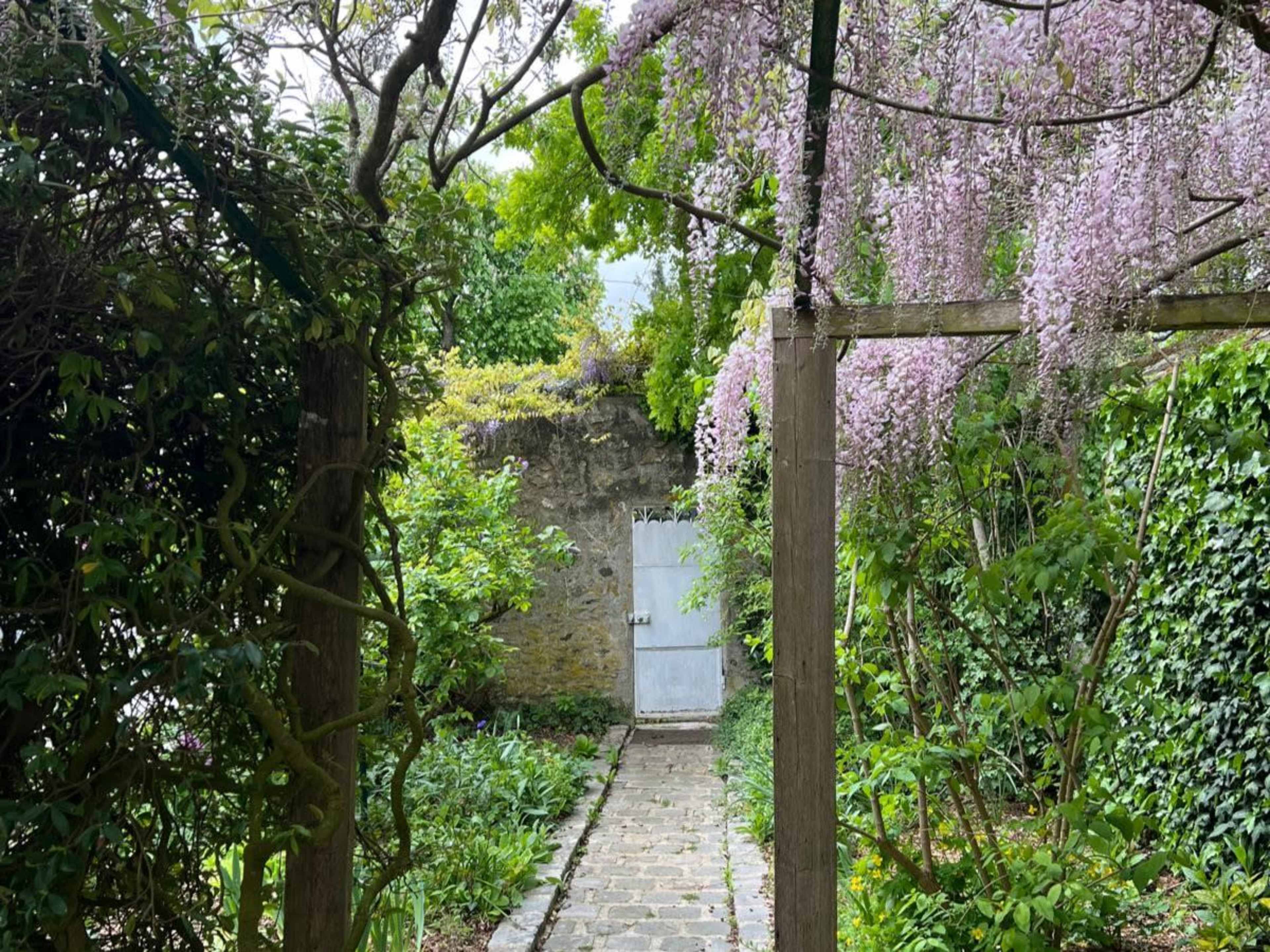 A stone pathway leads through a green garden, framed by wooden arches and flowering vines, towards a white door set into a stone wall.