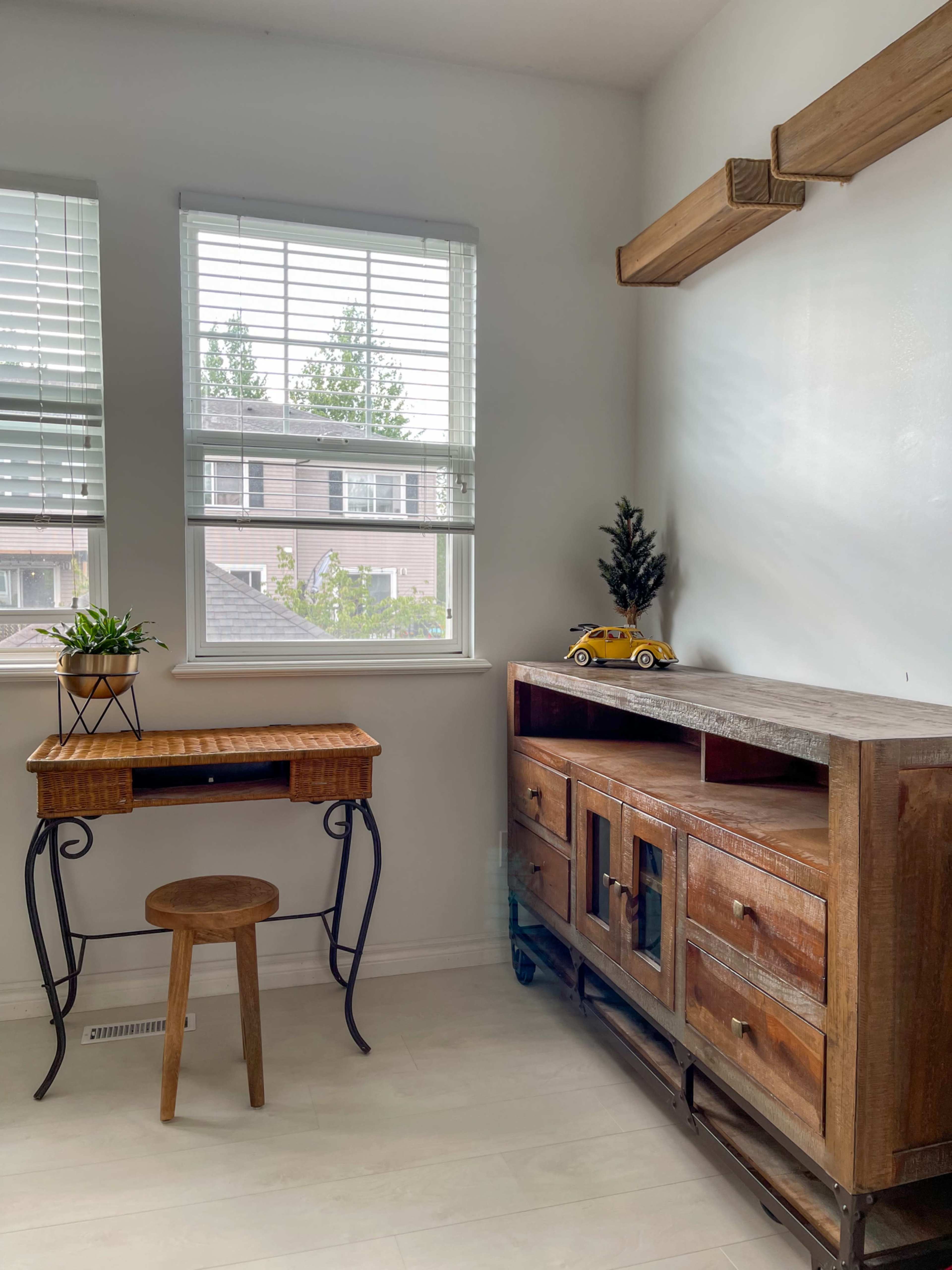 The image shows a small, bright workspace featuring a wooden desk with a stool, a decorative plant, and a wooden cabinet with drawers against a white wall, illuminated by natural light through windows with blinds.