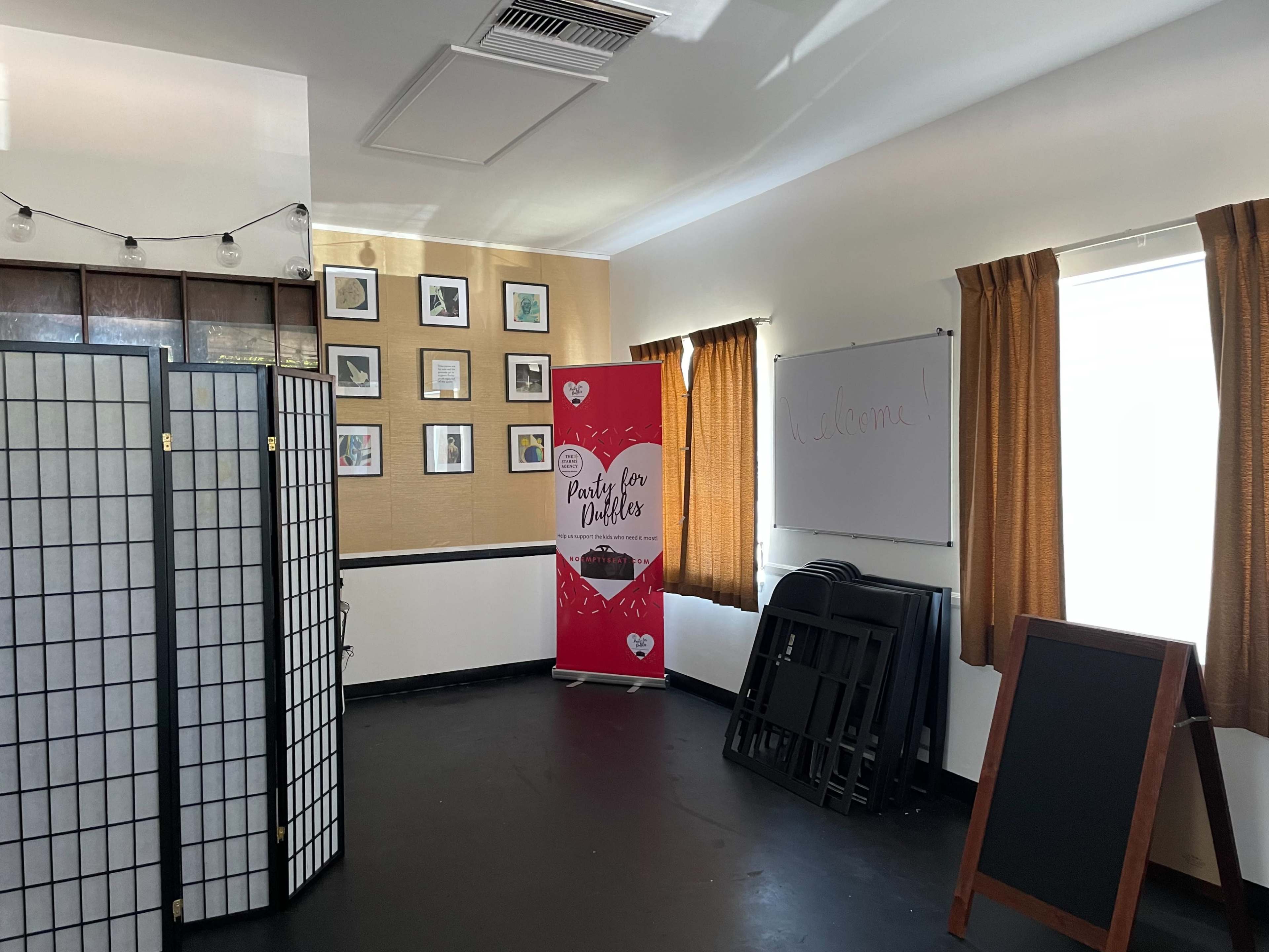 An interior room with a display banner, framed pictures on the wall, a whiteboard, and folding tables stacked against one side.