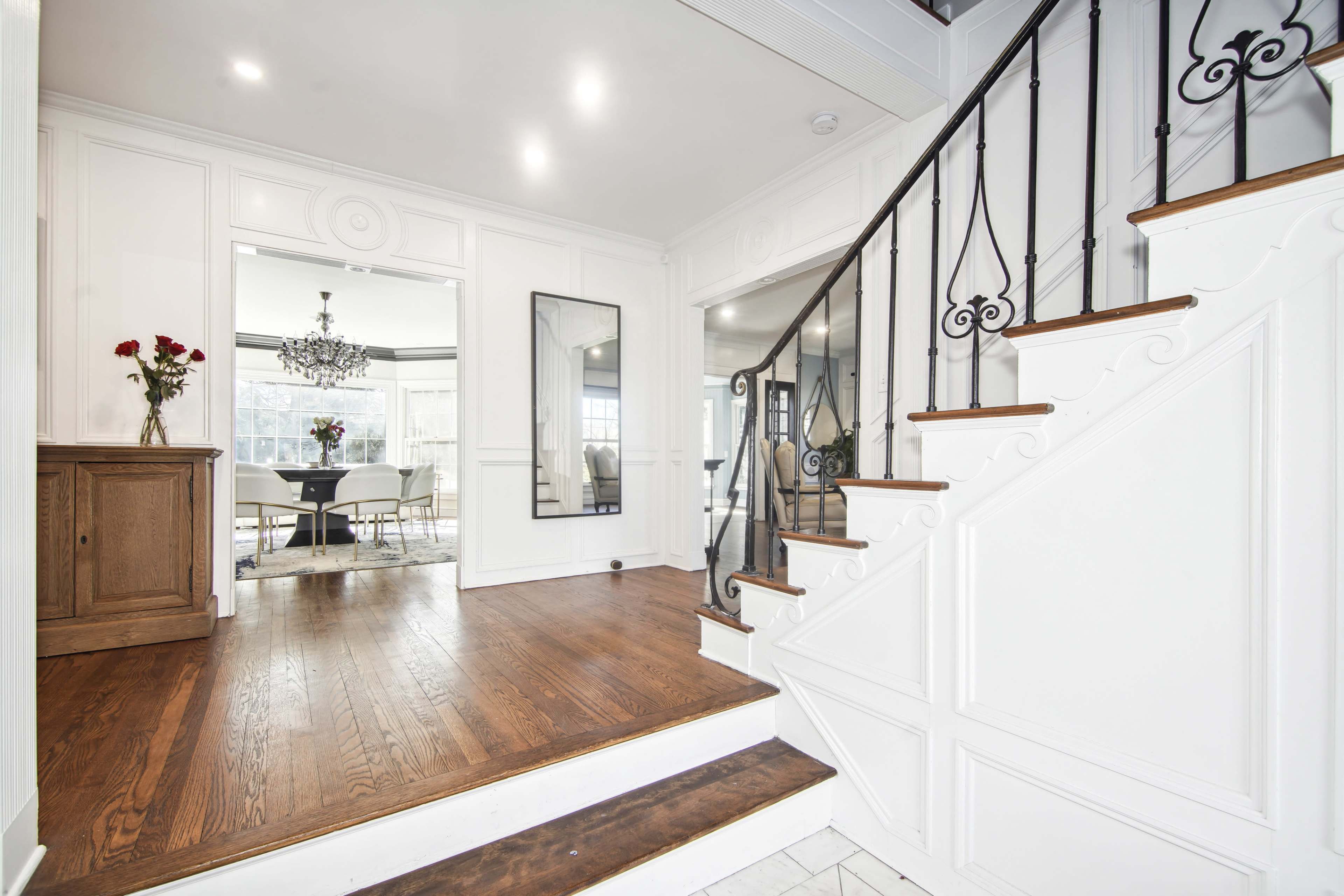 A well-lit entrance hall featuring a staircase with iron railings, a wooden console table, and a mirrored wall reflecting a dining area in the background.