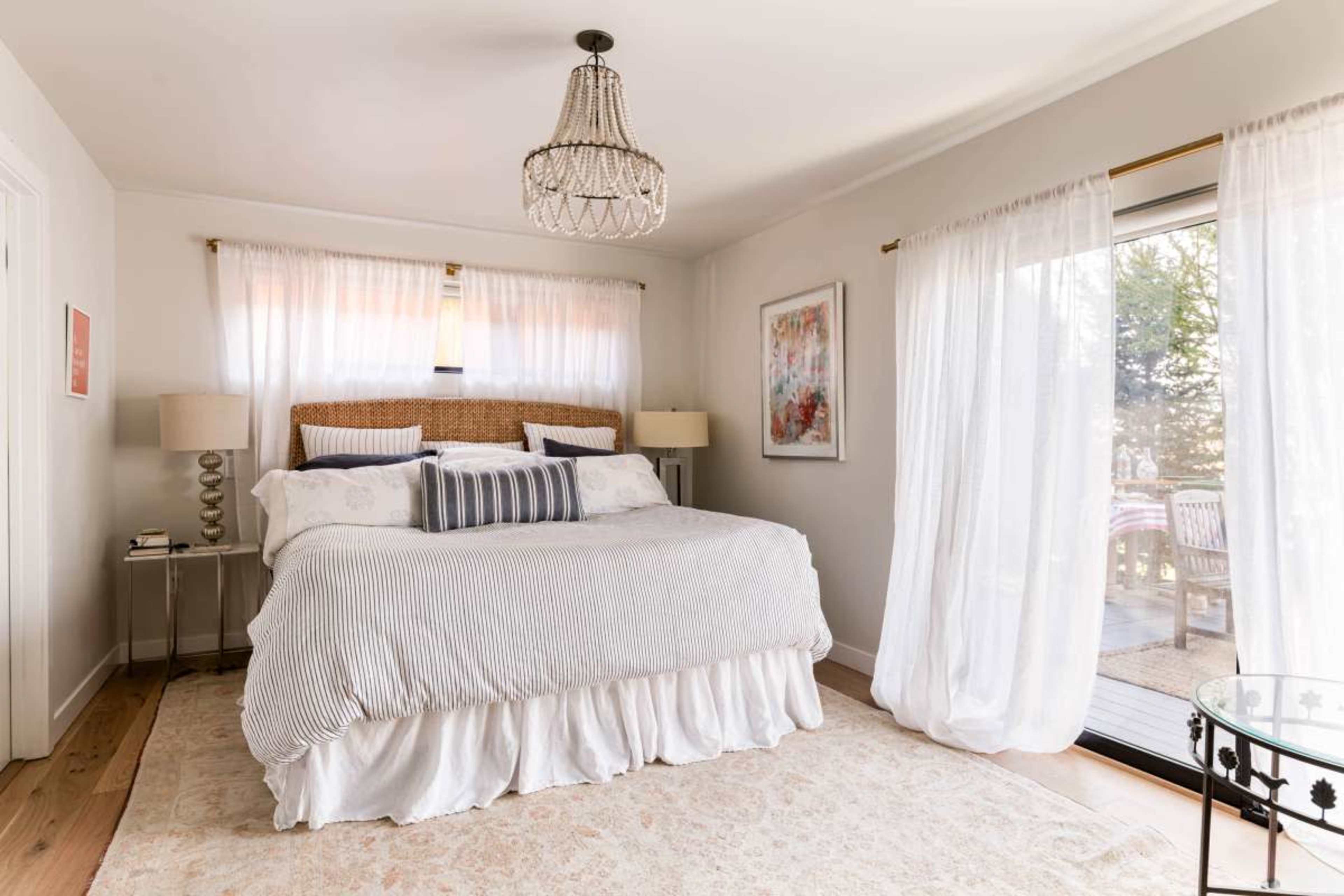 A spacious bedroom features a large bed with a light-colored comforter, two bedside lamps, and a sliding glass door that opens to an outdoor patio.