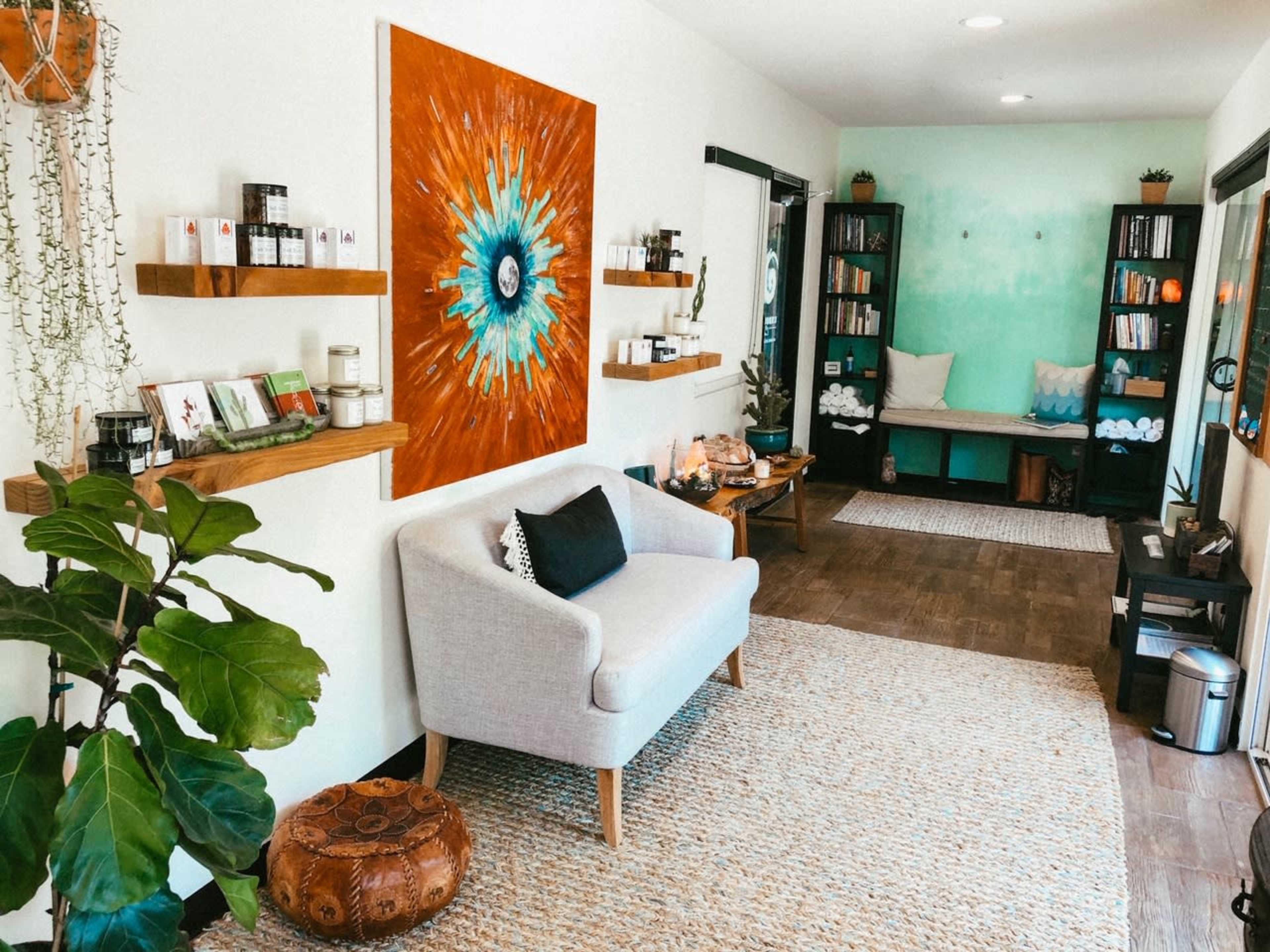 A cozy wellness space featuring a grey armchair, a large vibrant artwork, wooden shelves lined with jars and books, and a seating area with plants and decorative elements.