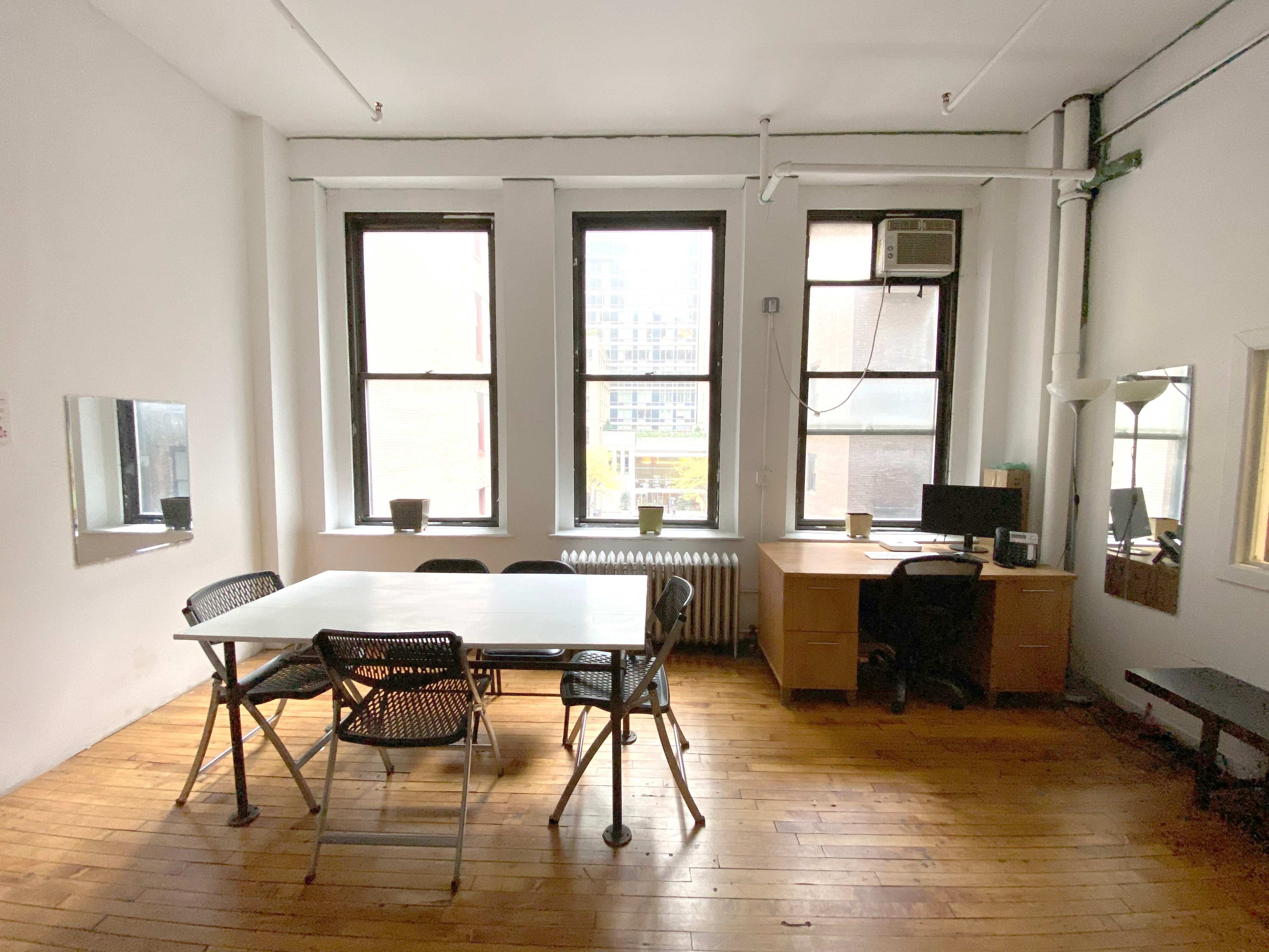 A bright, empty room with wooden floors, a large table surrounded by chairs, a desk with a computer, and windows letting in natural light.