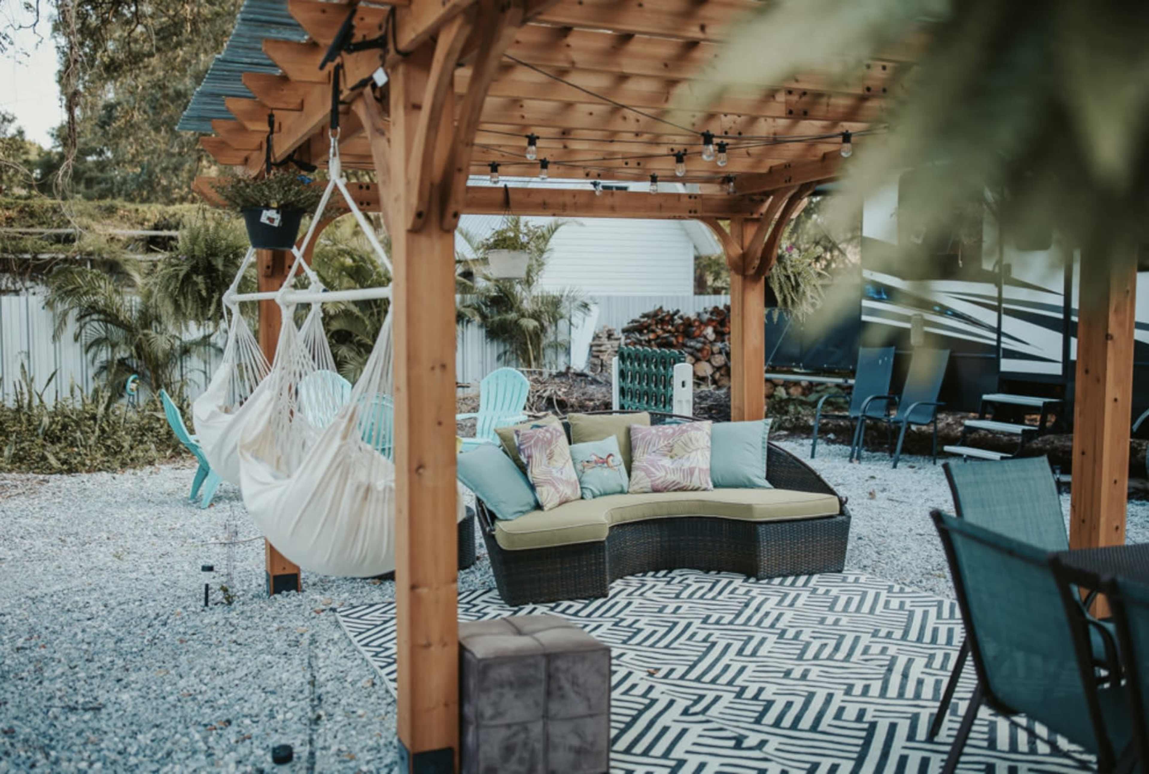 A wooden pergola with a hammock and a sectional sofa is set on a gravel patio with decorative plants and outdoor furniture.