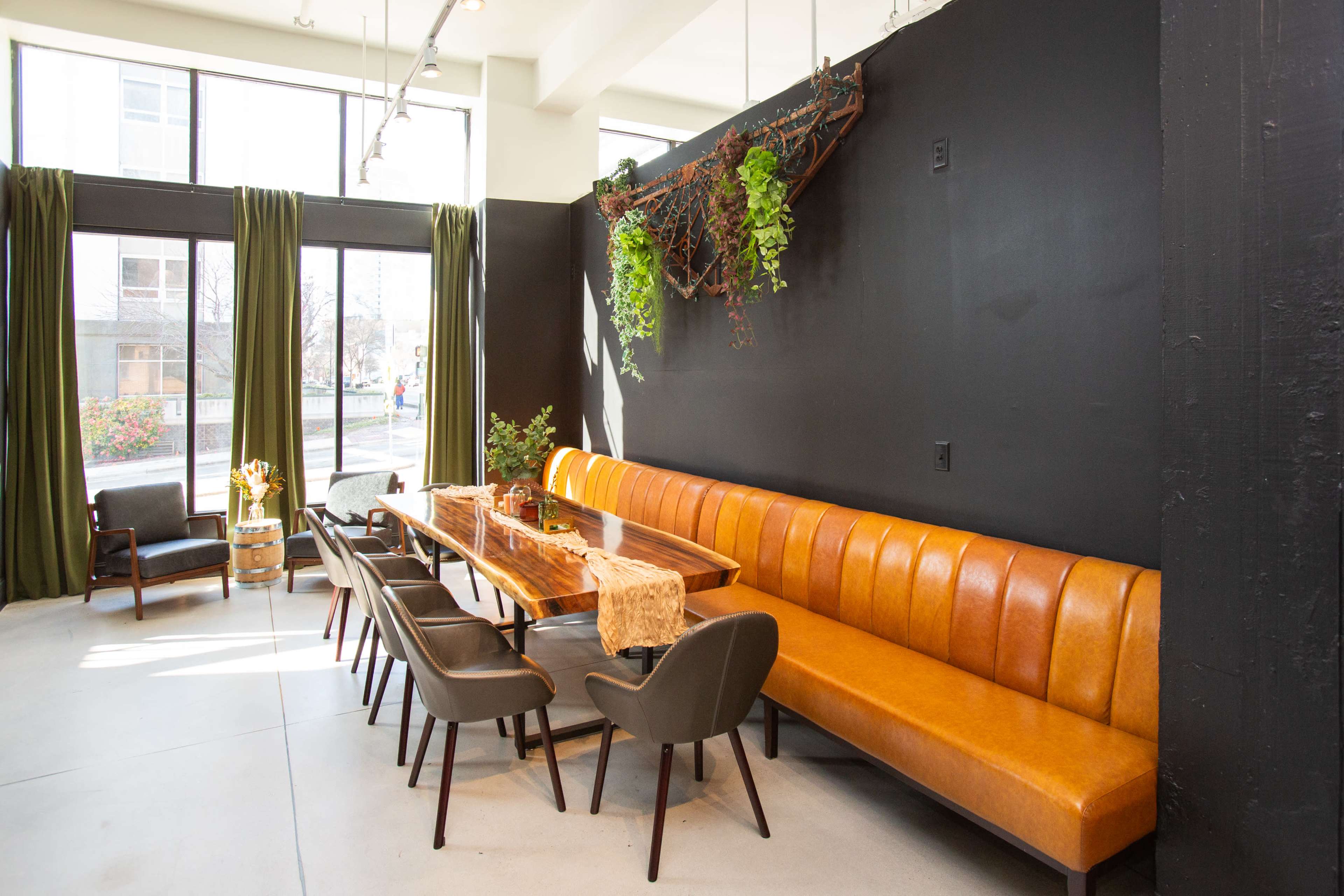 The image shows a modern dining area with a long wooden table surrounded by black chairs and a brown leather bench, situated near large windows with green curtains.