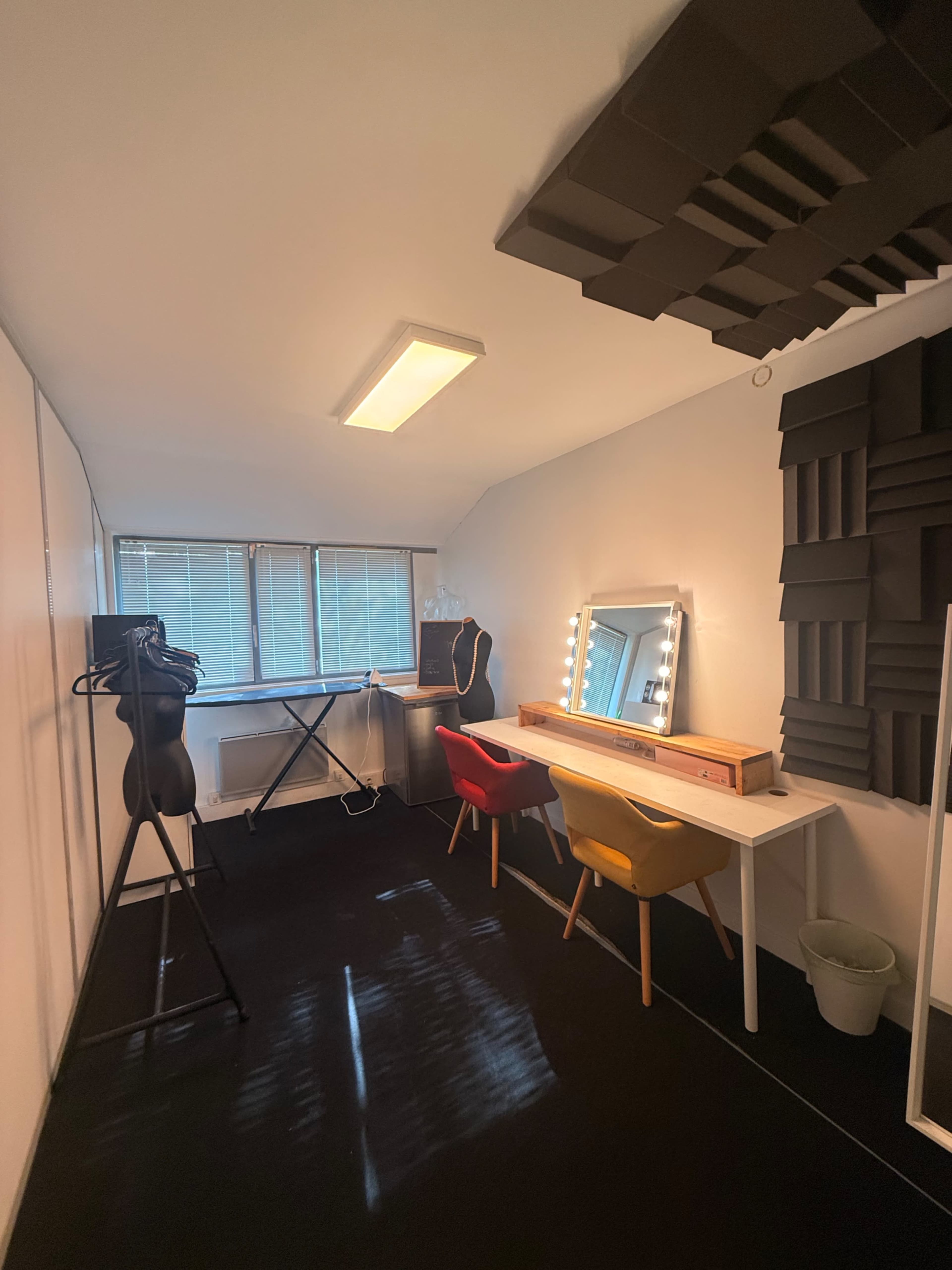 The room features a minimalist design with a piano, a large mirror with lights, two colorful chairs, and soundproofing panels on the walls.