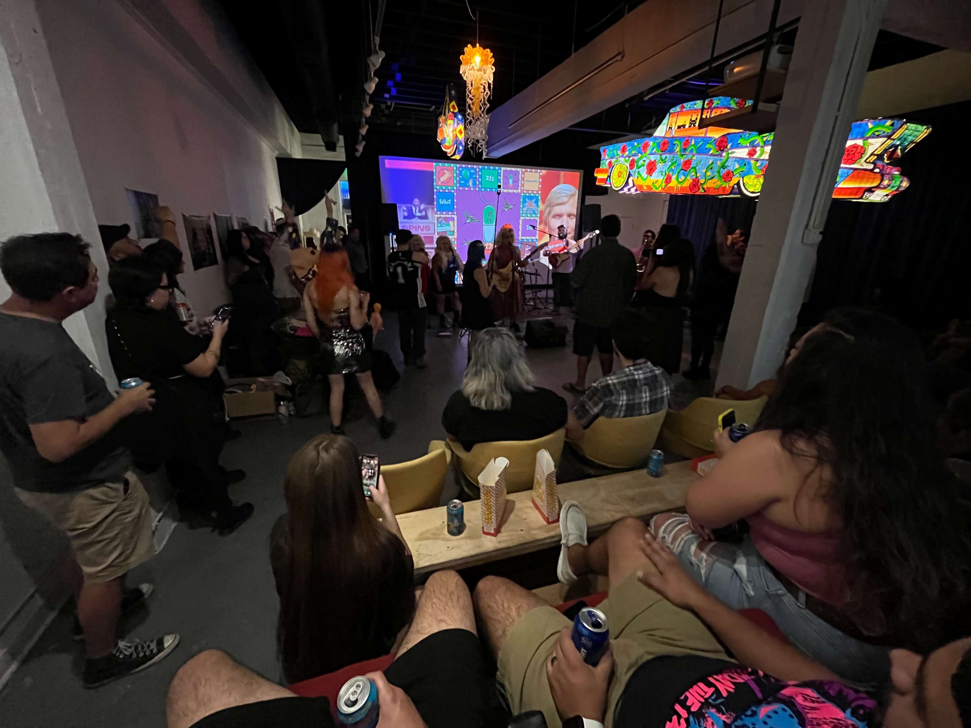 A crowd gathers in a dimly lit venue, watching a performance on a screen while some attendees take photos and videos.