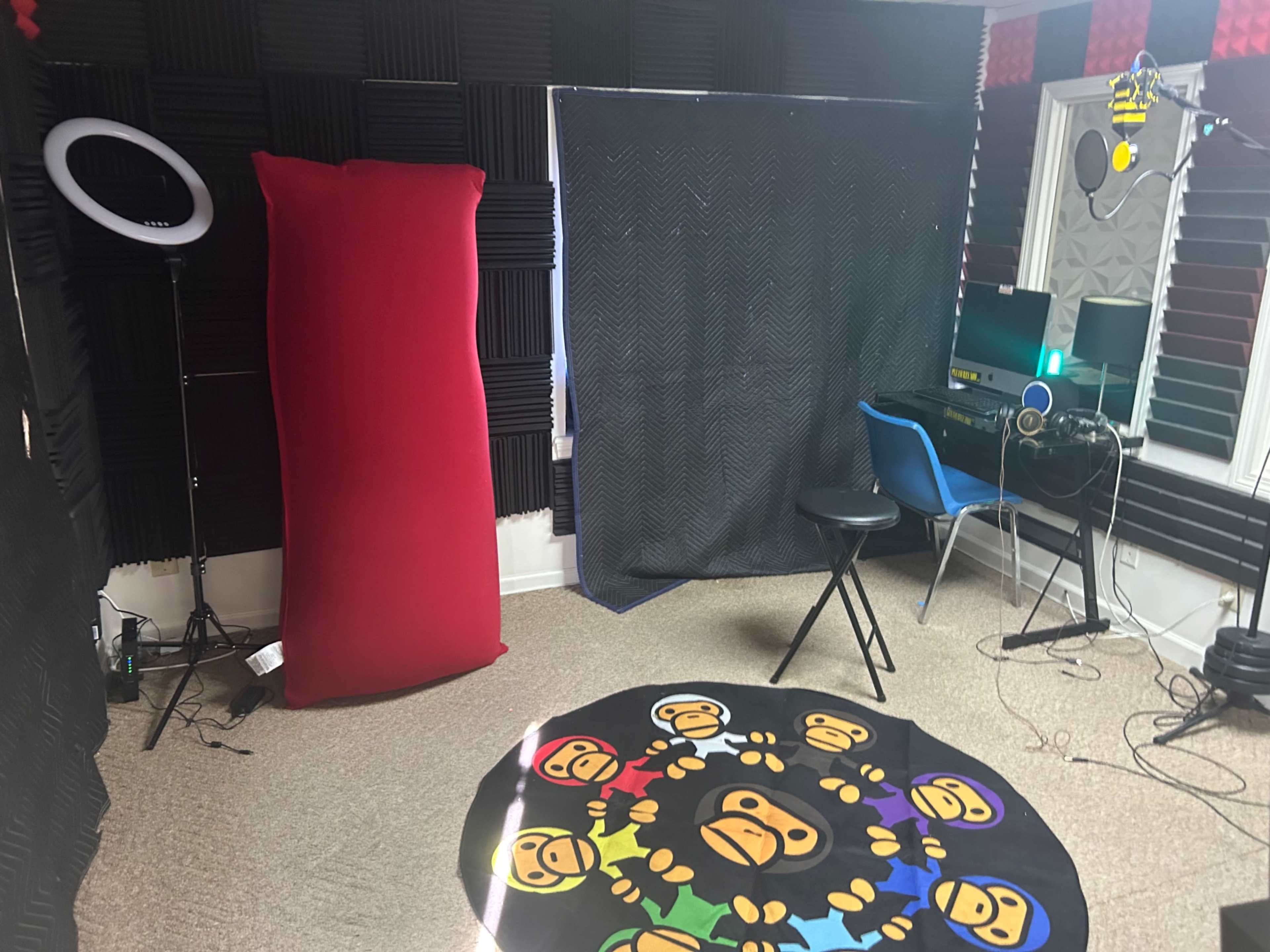 A recording studio with soundproofing panels, a blue chair, a computer setup, a black stool, and a circular rug featuring cartoon monkeys.