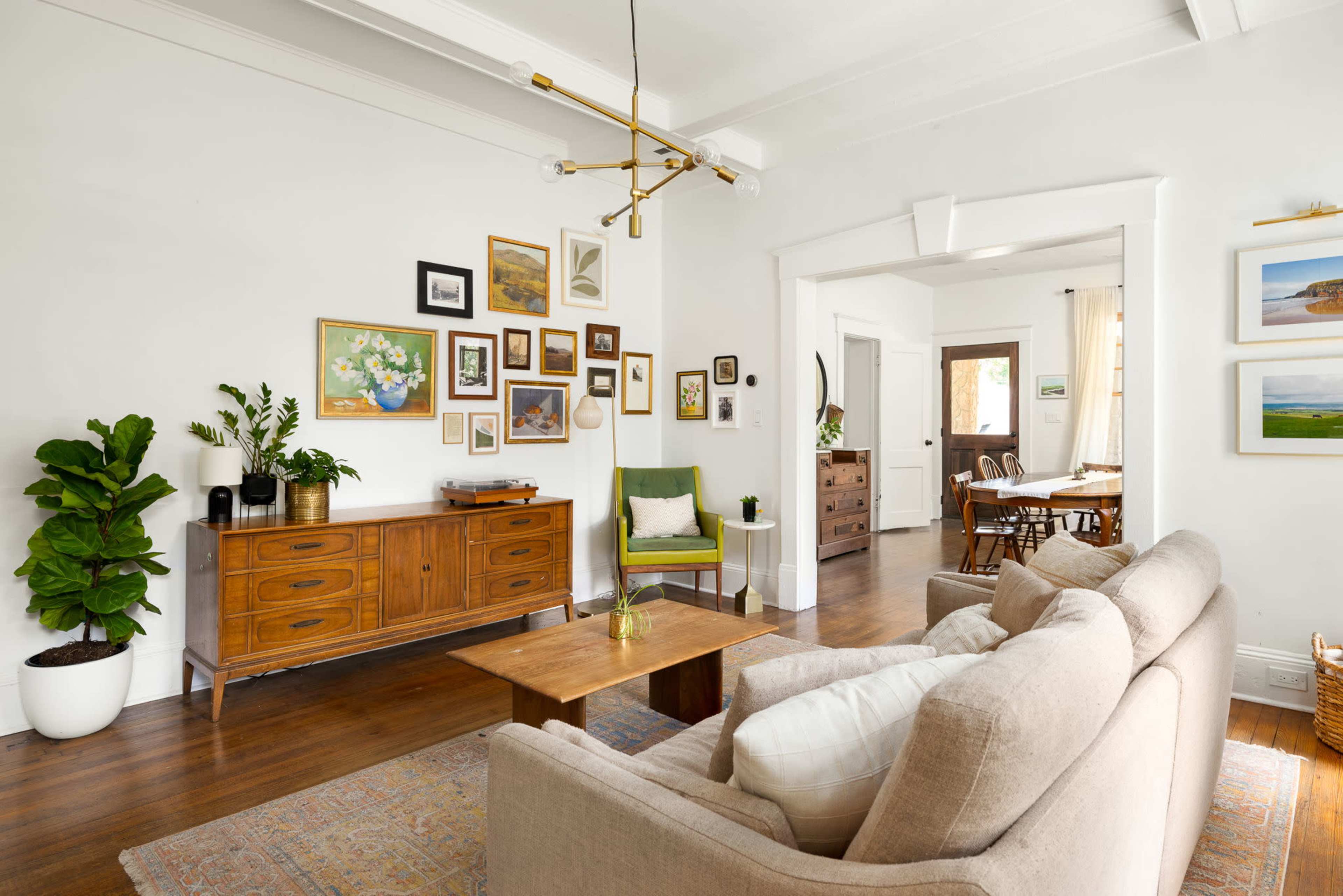A cozy living room features a beige sofa, a wooden coffee table, and a sideboard adorned with framed artwork and plants, with an adjacent dining area visible in the background.