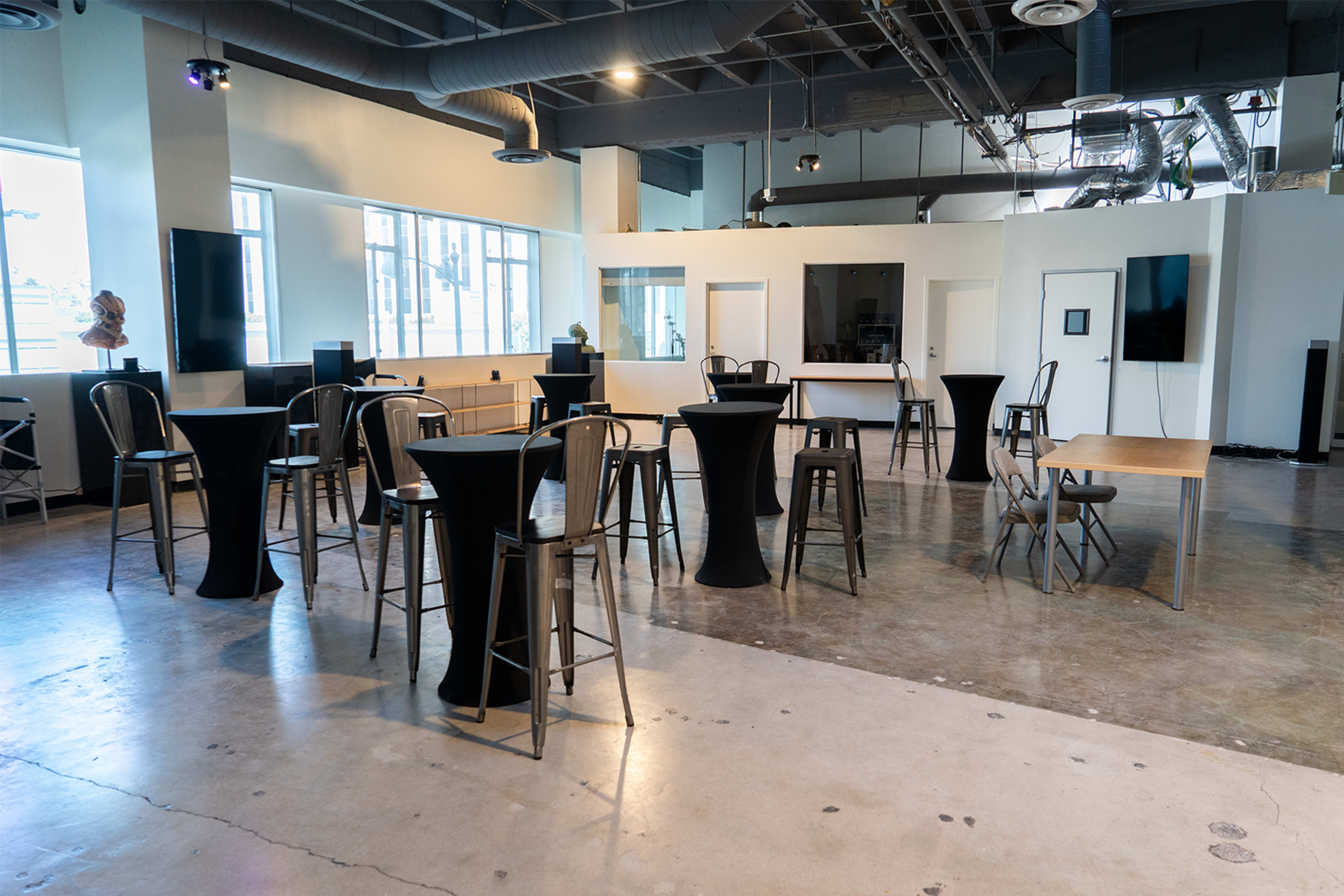 A spacious event room featuring several high tables with black tablecloths and chairs arranged around a polished floor.