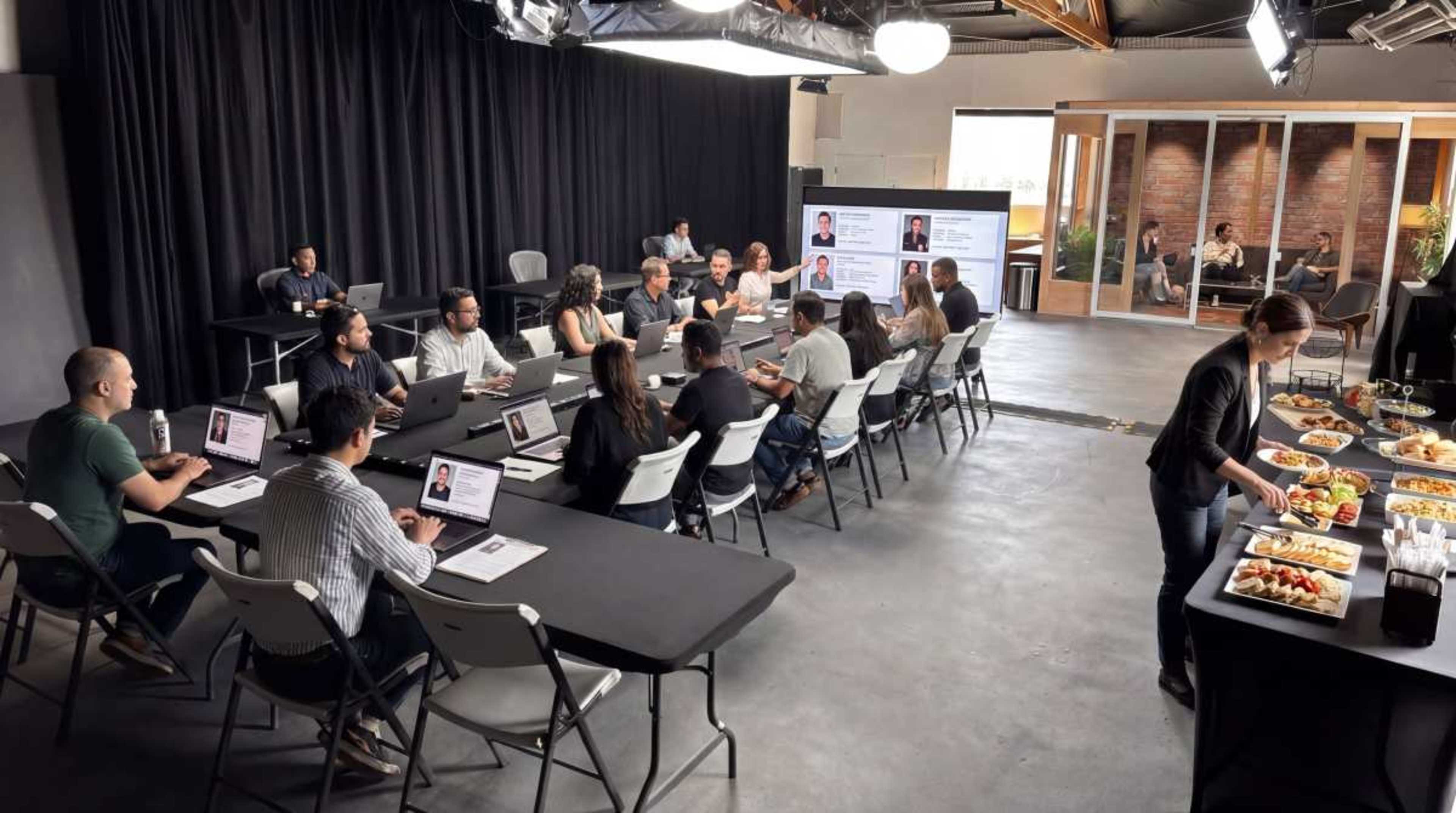 A diverse group of people sits around a long table with laptops, while a woman serves food from a buffet in a well-lit meeting space.