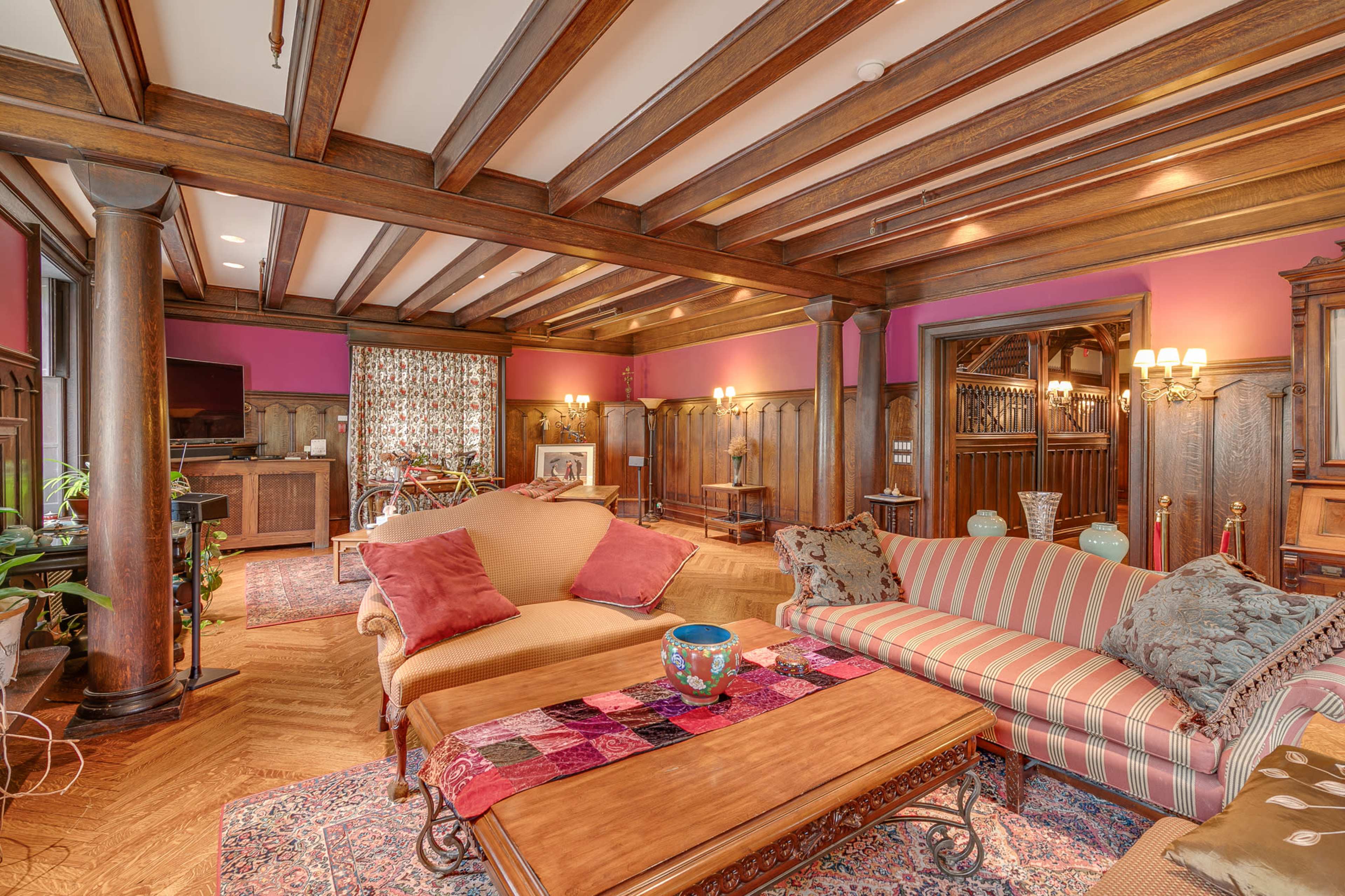 The room features wooden beams on the ceiling, a mix of furniture including a striped sofa and a coffee table, and decor elements such as plants and a patterned area rug.