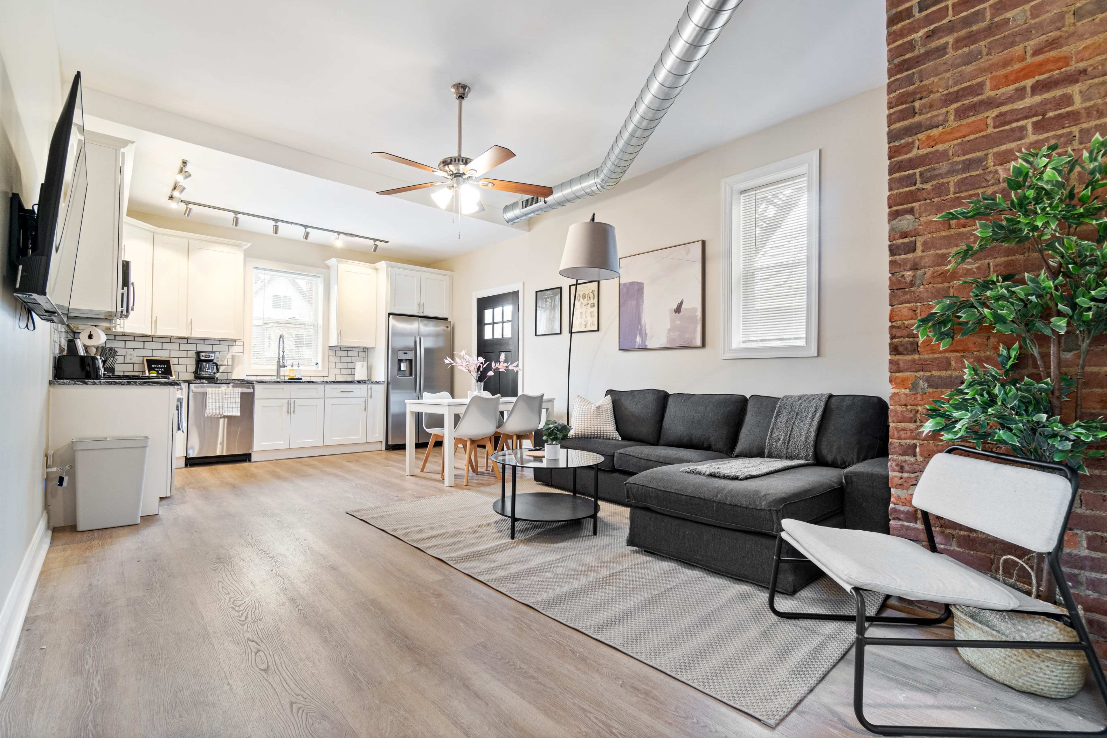 The image shows a modern living space featuring a sectional couch, a dining area with a table, and a kitchen with white cabinetry and stainless steel appliances.