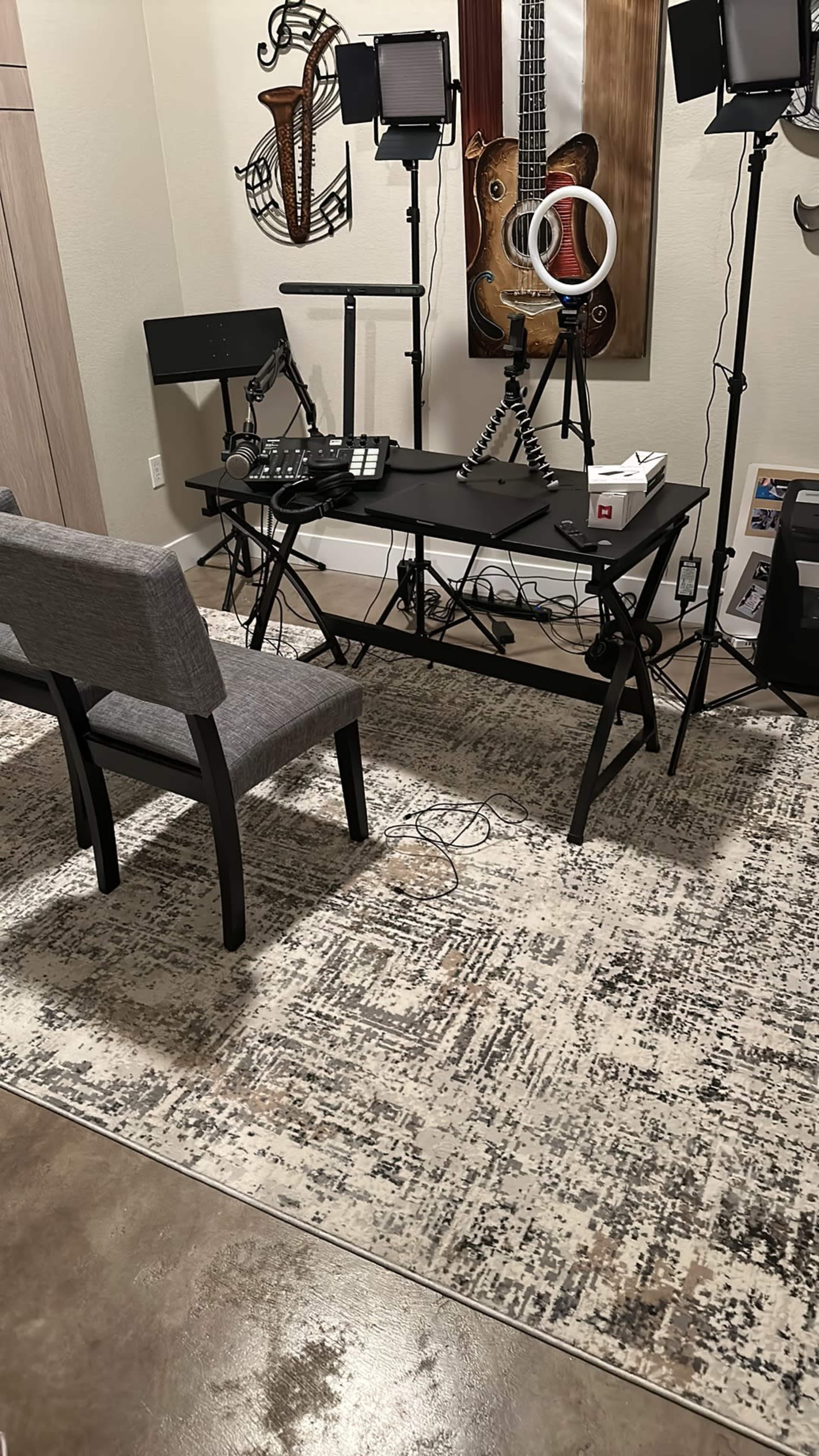 The image shows a minimalist workspace featuring a black desk with equipment, a gray chair, and several lights, set against a muted wall decorated with musical instruments.