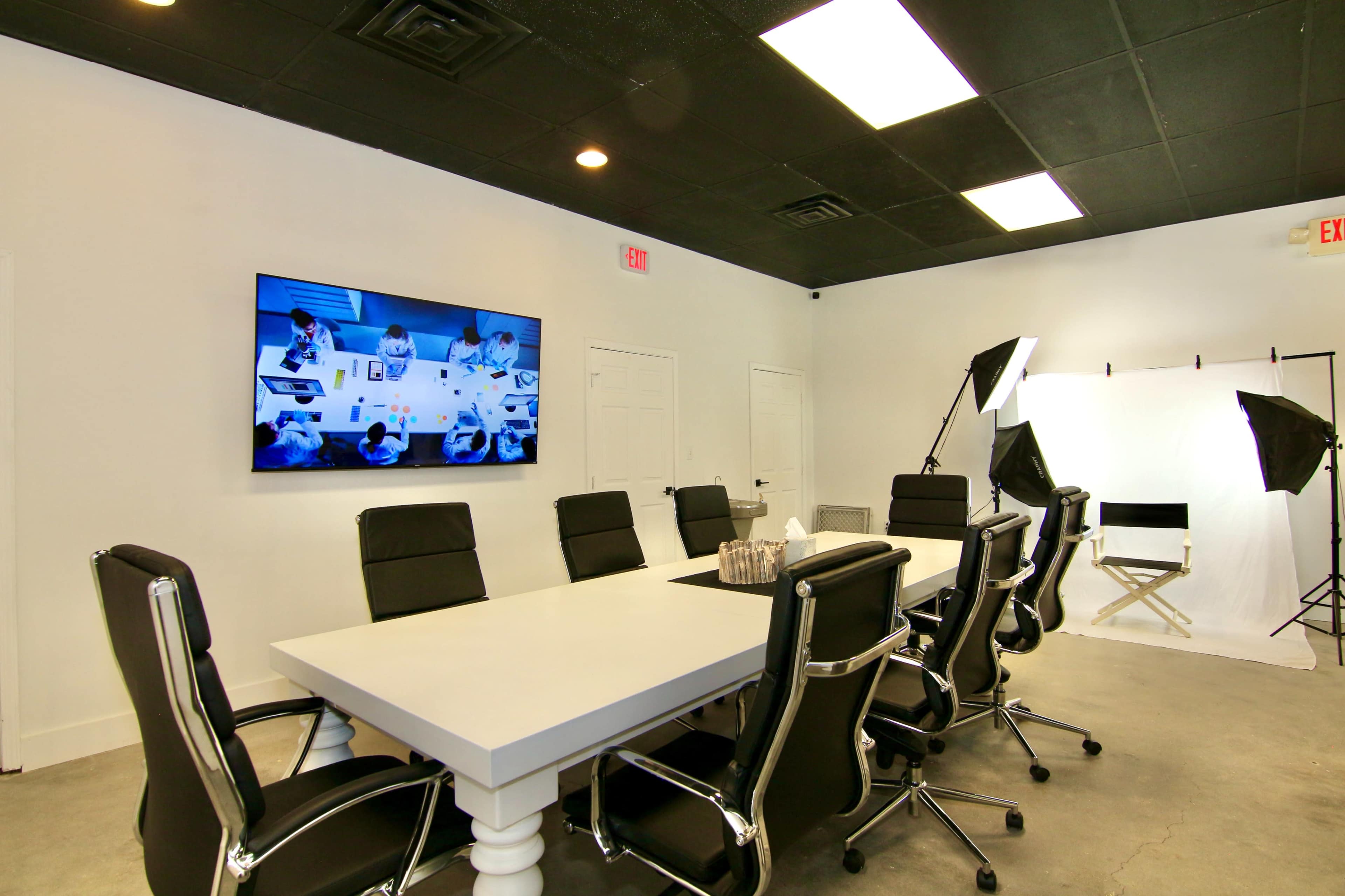 A conference room features a long white table with black chairs, a large screen displaying a meeting, and professional lighting equipment in the corner.